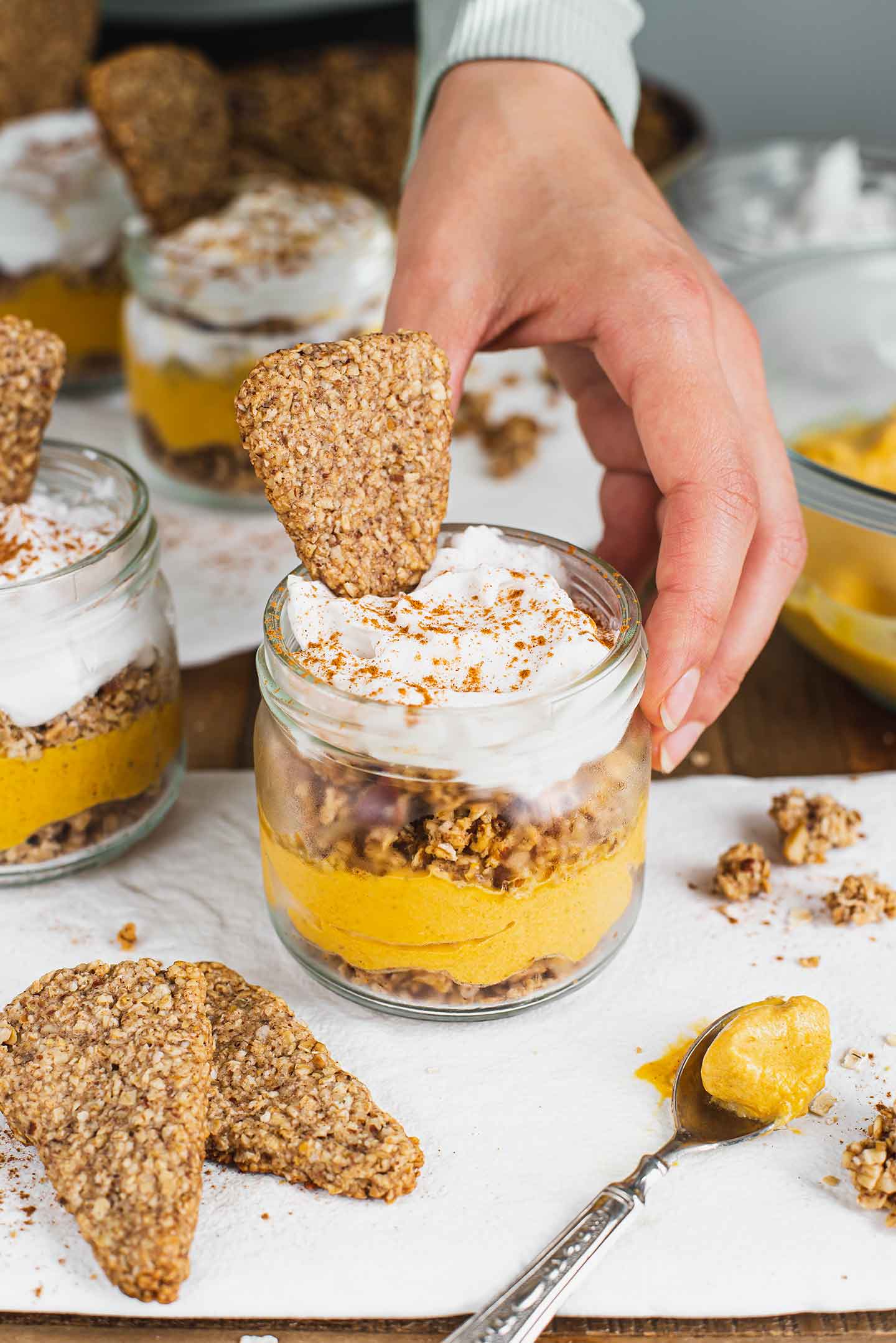 Side view of a hand placing a personal pumpkin pie parfait on a white tray. Creamy pumpkin mousse is visible between two layers of granola. Fluffy whipped cream tops it off and a pie shaped cookie sticks up from the whip.