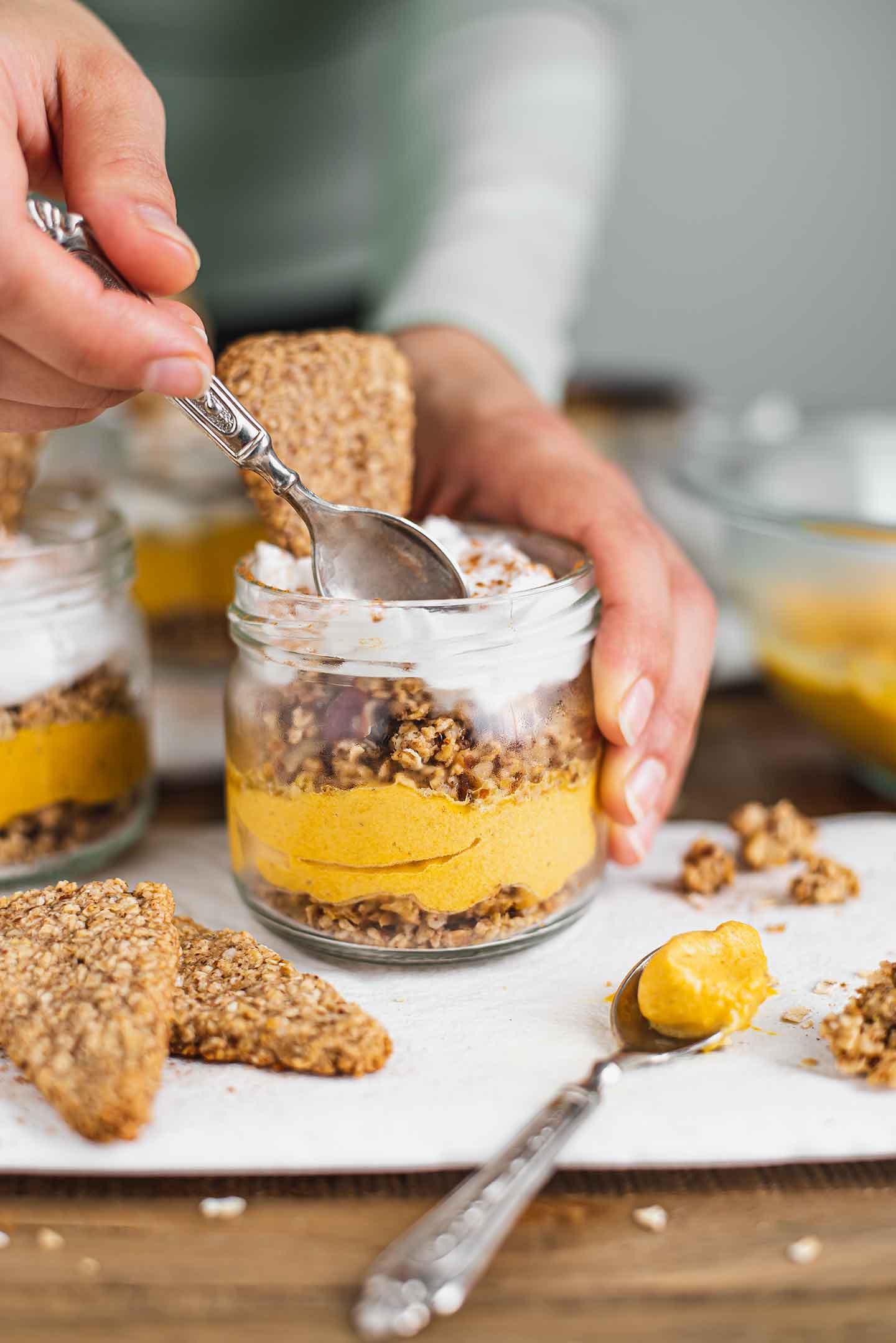 Side view of a hand dipping a spoon into a parfait. Pie crust cookies and a dollop of pumpkin mousse lay beside the jar.