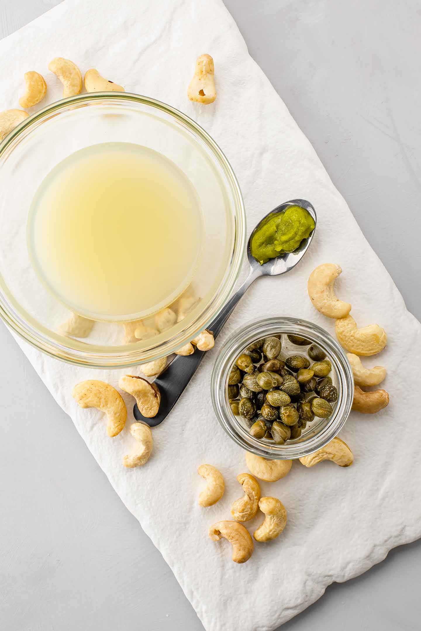 Lemon juice in a bowl, wasabi on a teaspoon, capers in a small jar and scattered raw cashews are displayed on a white slab