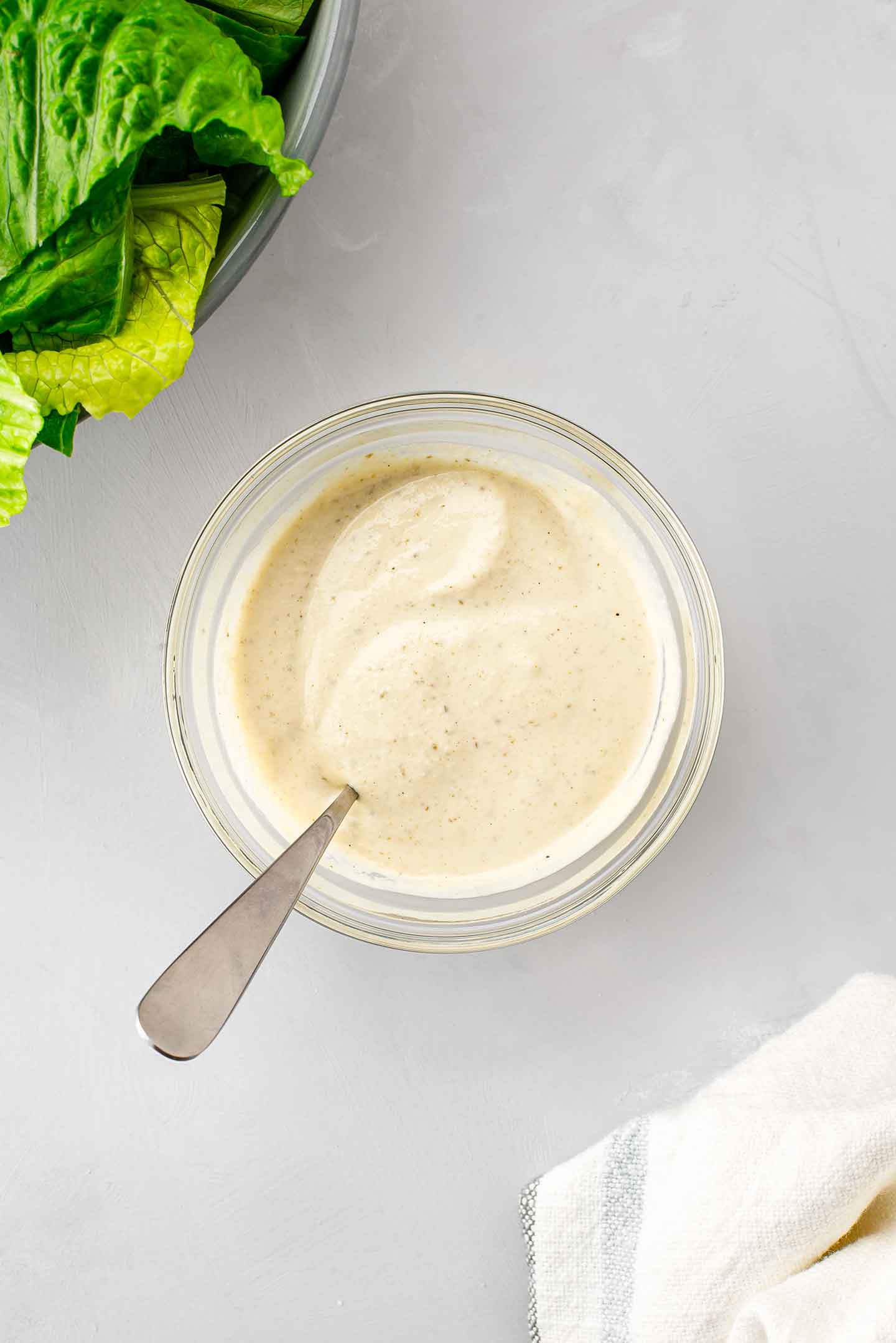 Top down view of creamy caesar dressing in a bowl with a spoon and romaine lettuce leaves in the corner