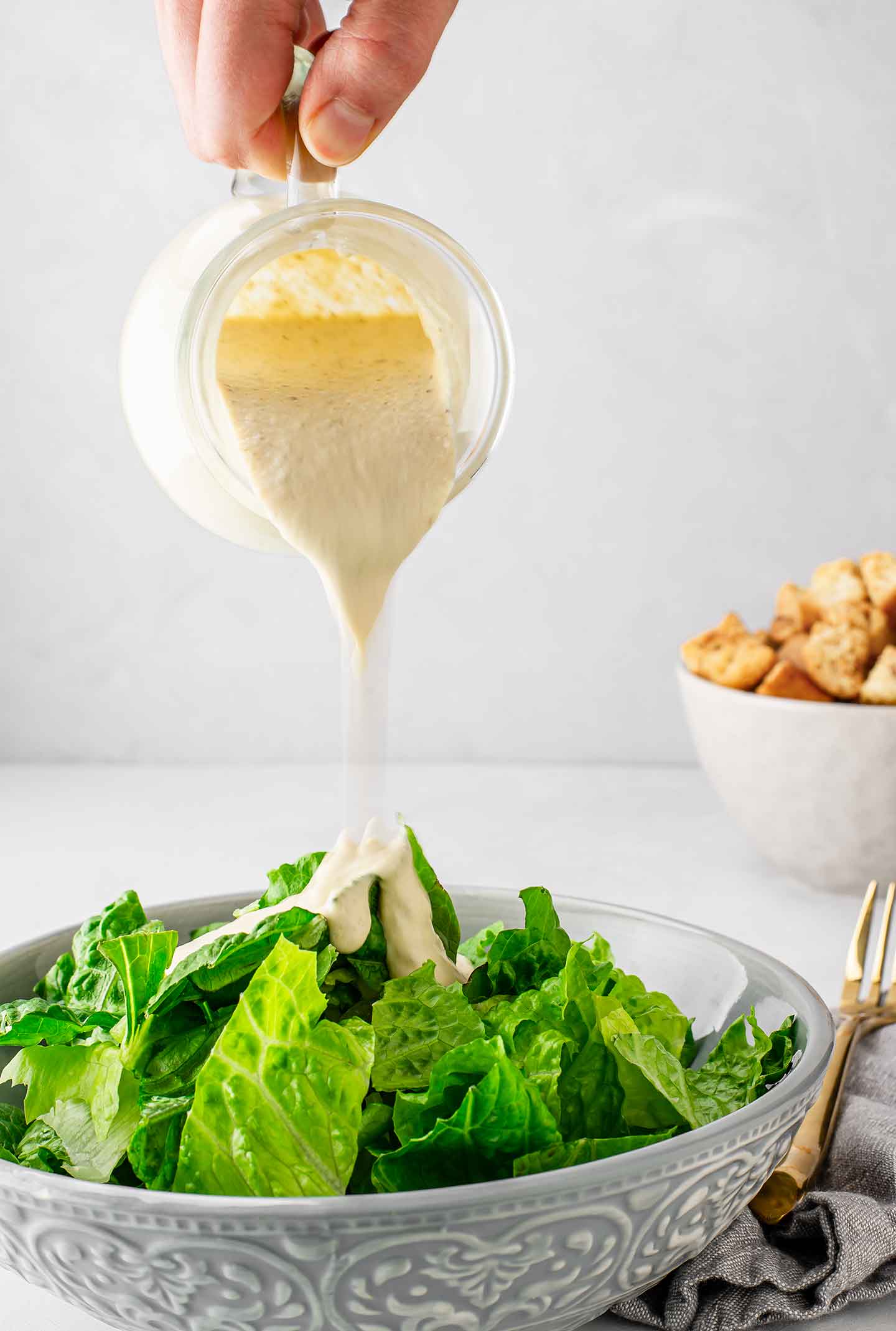 Zingy caesar dressing being poured from a glass jar over a bowl of romaine lettuce with croutons in a bowl in the background