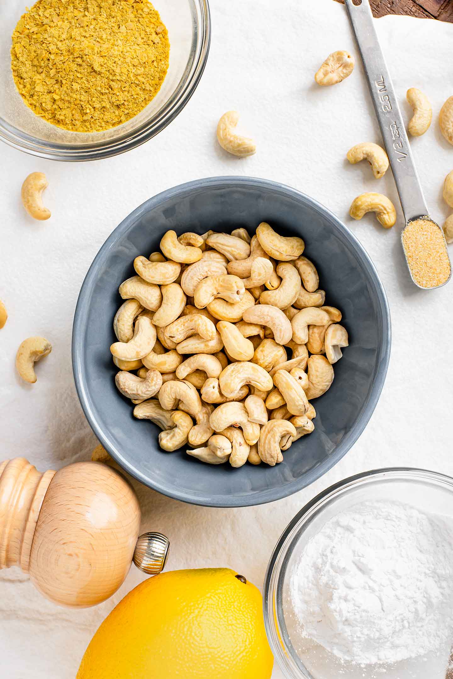 Top down view of ingredients. A bowl of raw cashews is in the centre of a white tray with nutritional yeast, garlic powder, a salt mill, a lemon, and tapioca flour surrounding.