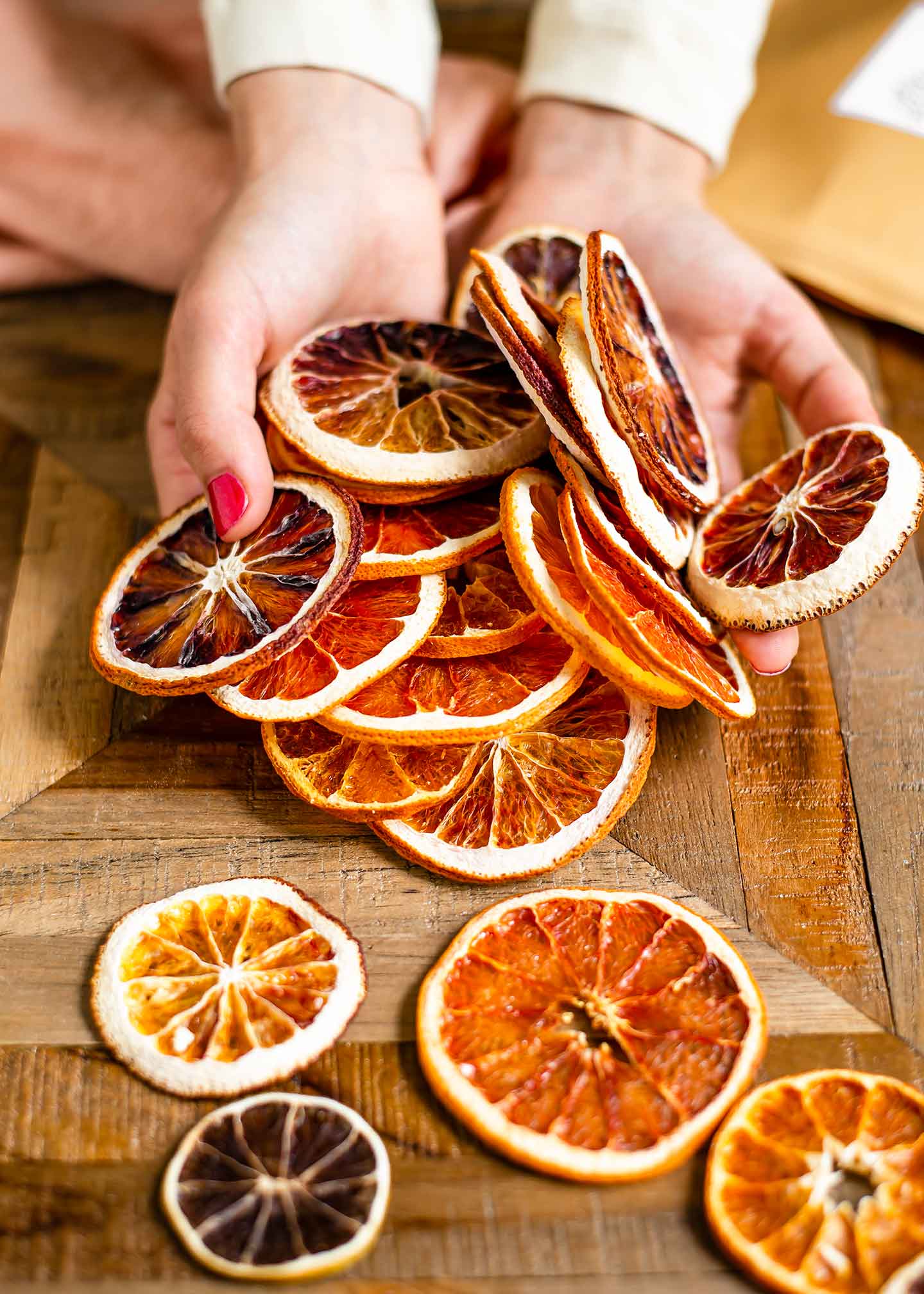 Top down view of sliced and dried citrus fruit spilling out of two hands and on to a wooden table. The dried fruit are different shades of red and orange and varying sizes.