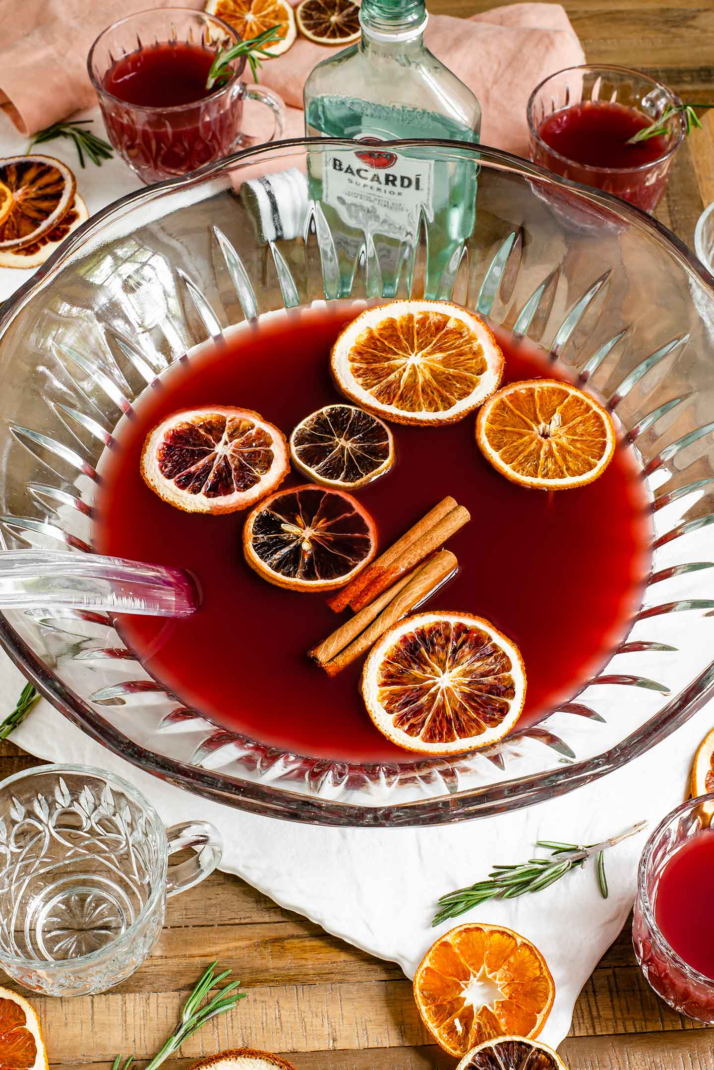 Top down view of a deep red pomegranate rum punch in a punch bowl. The drink is garnished with dried citrus fruit and cinnamon sticks.