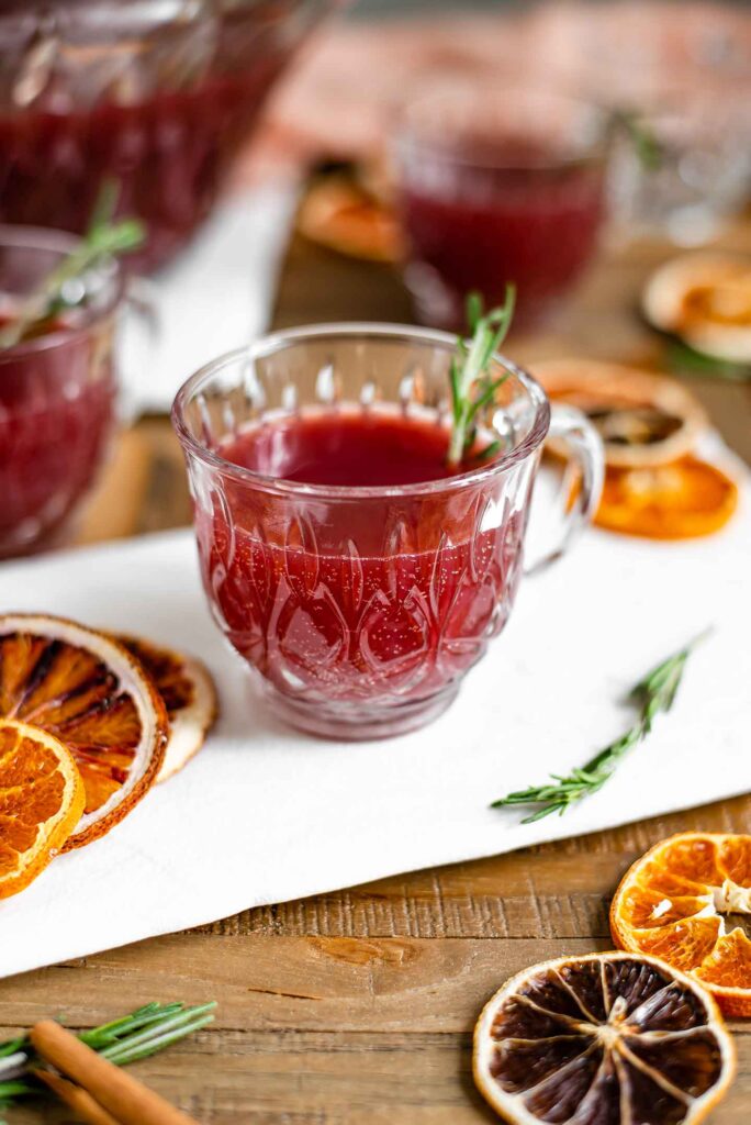 Side view of a small punch glass on a white tray filled with a deep red bubbly punch. A sprig of rosemary garnishes the glass and dried citrus fruit is scattered on the tray.