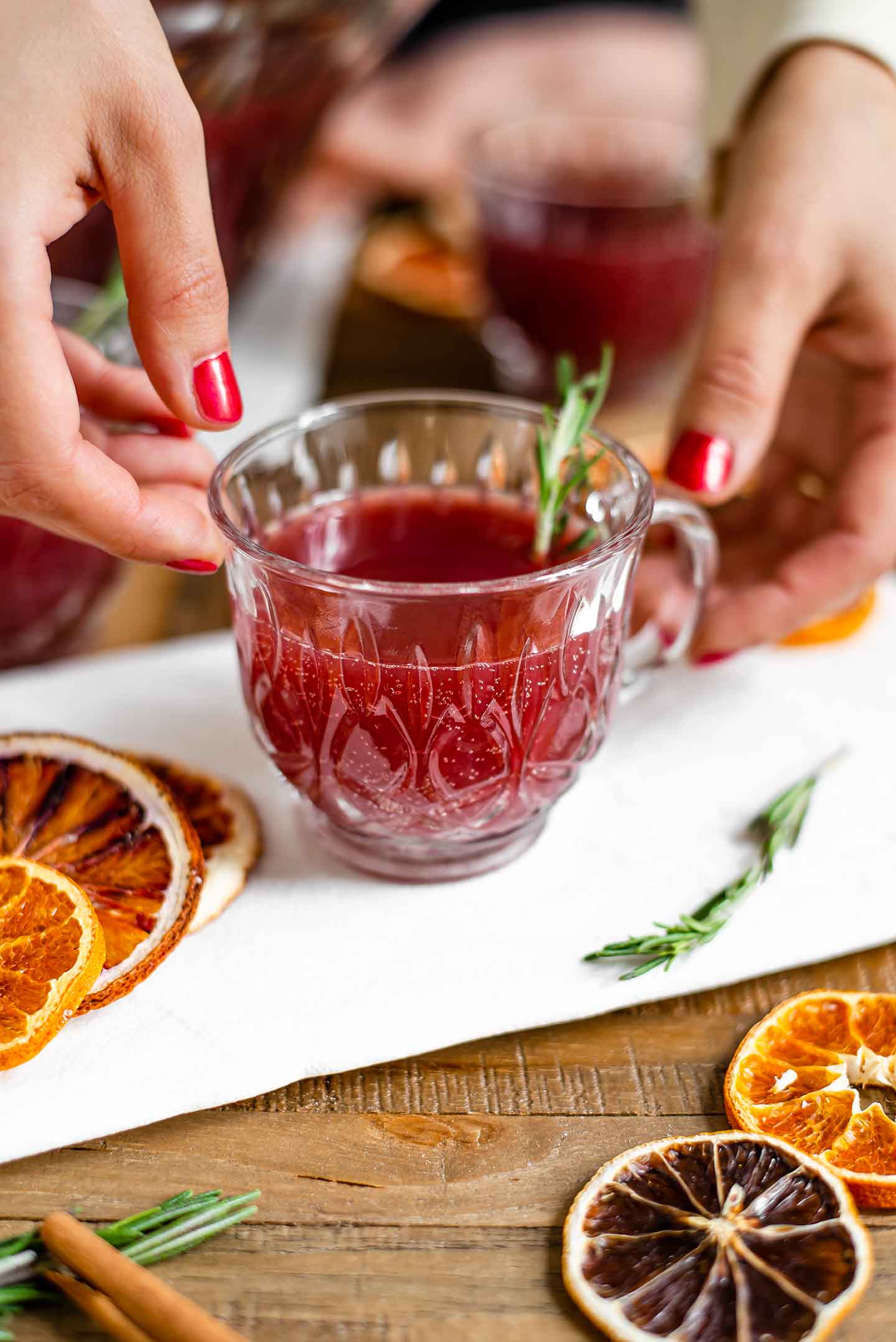 Side view of hands reaching for a small punch glass filled with pomegranate rum punch. Small bubbles can be seen and the glass is garnished with a sprig of rosemary.