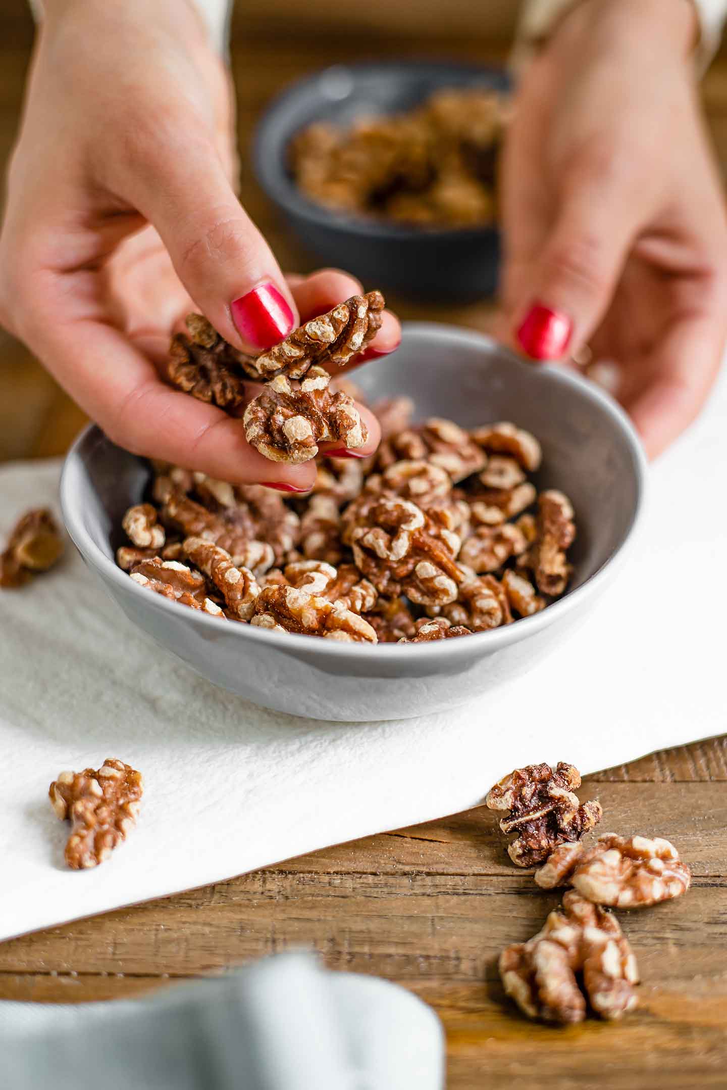 Side view of a hand holding toasted walnuts. Raw walnuts are in the background and loose walnuts are scattered around on the wooden table.