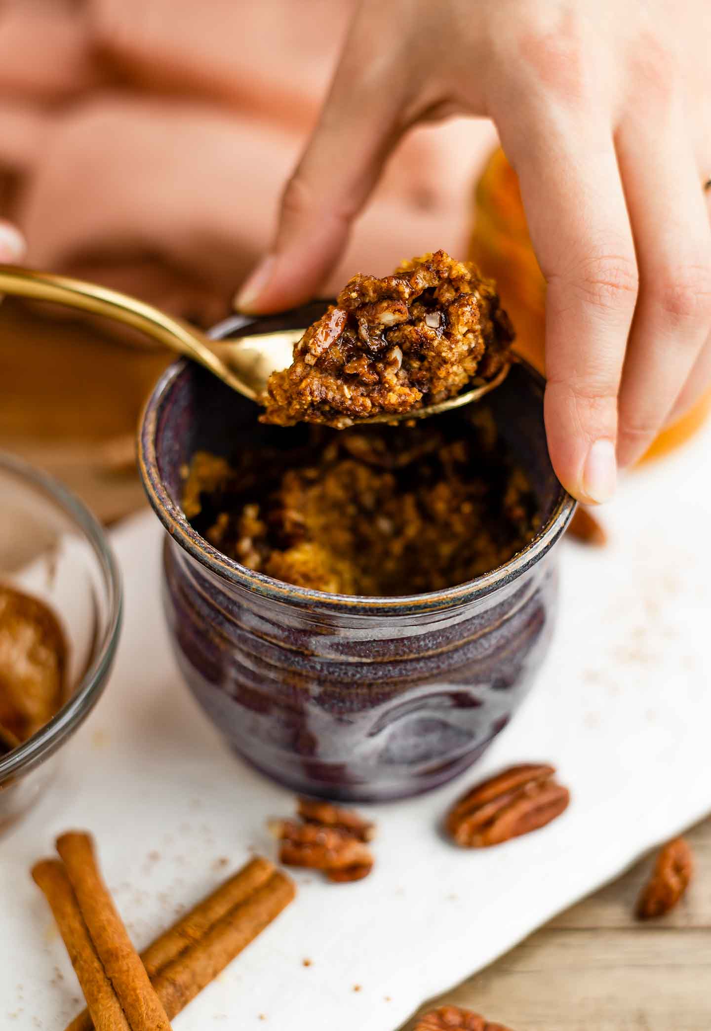 Side view of a hand holding a scoop of warm pumpkin spice mug cake on a spoon. Crushed pecans and a maple cinnamon drizzle are visible.