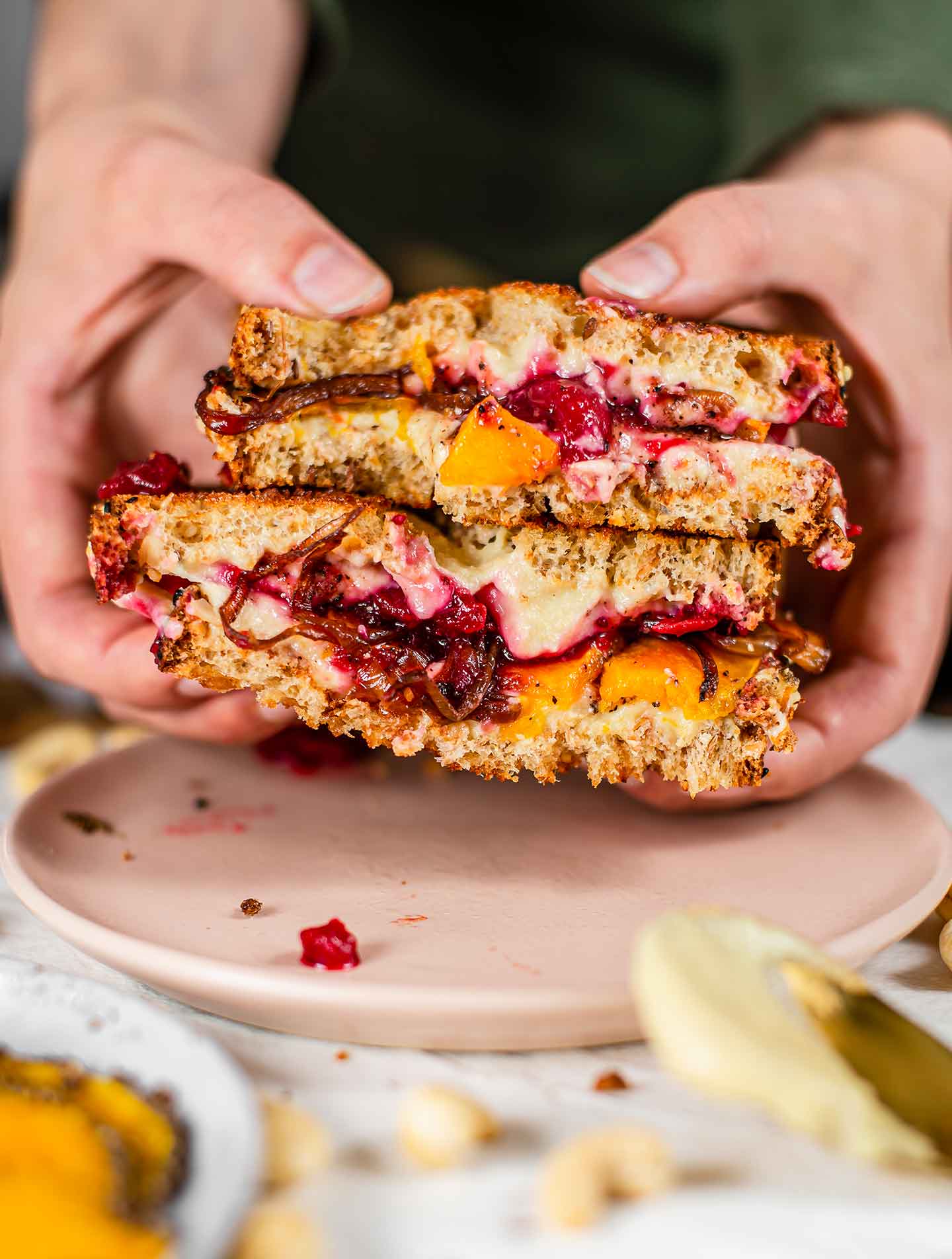 Side view of hands lifting a holiday squash sandwich from a plate. The sandwich has red cranberry sauce, white and gooey mozzarella, and orange roasted squash.