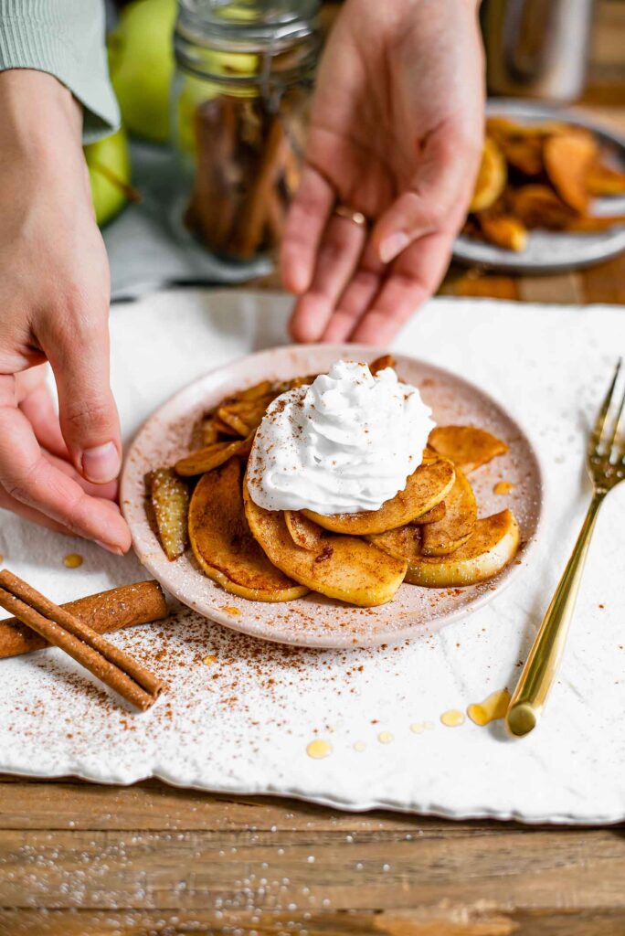 Coconut whip cream sits atop warm, tender apple slices coated in cinnamon and a dusting of icing sugar on a pink saucer. Two hand gently hold the edges of the saucer. Apples, cinnamon sticks and another plate of warm cinnamon fried apples sit in the background. Cinnamon, maple syrup, and cinnamon is scattered around the plate.