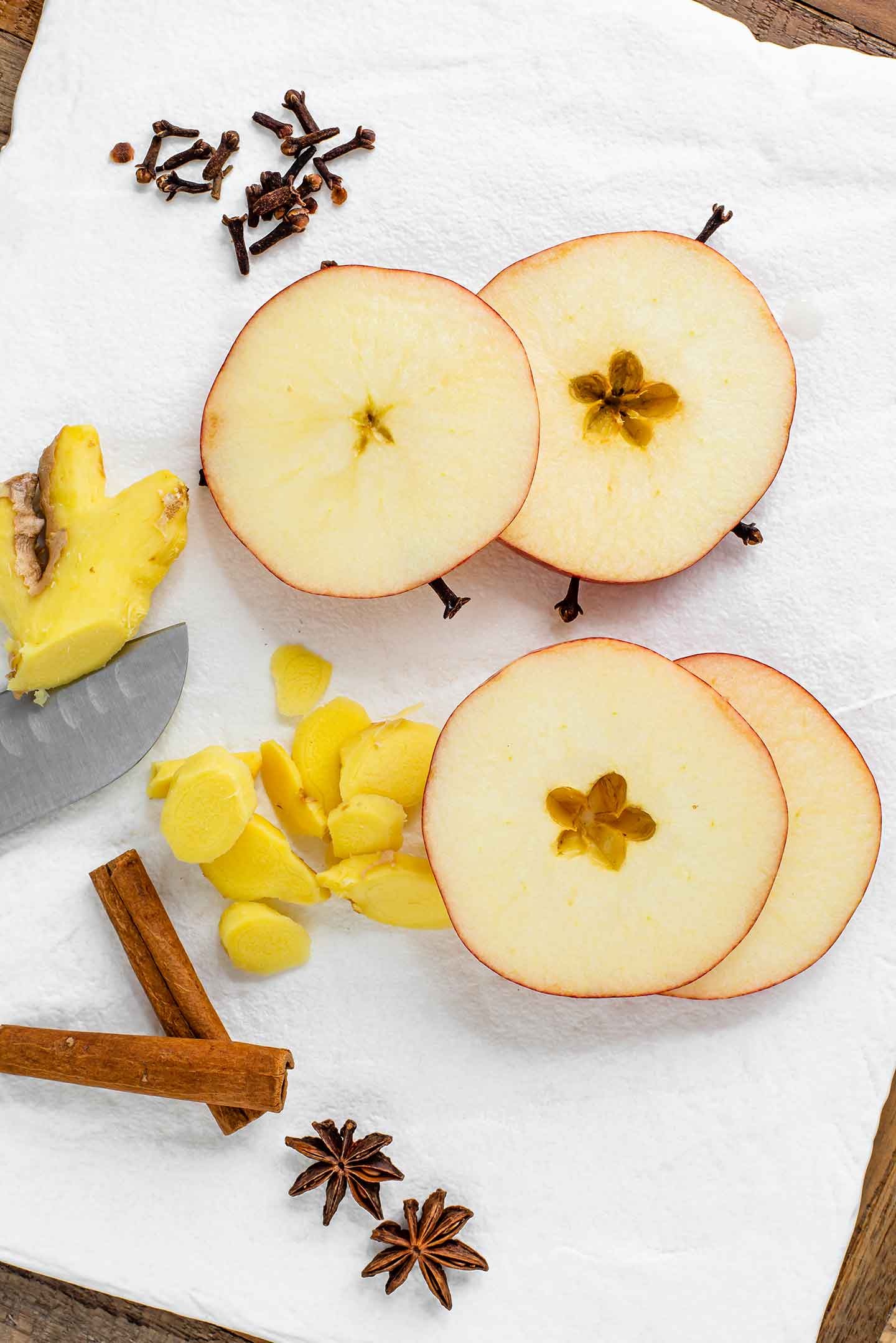 Top down view of sliced ginger, cinnamon sticks, star anise, and whole clove. An apple sliced into 4 rings and some of the slices have been poked with the cloves. 