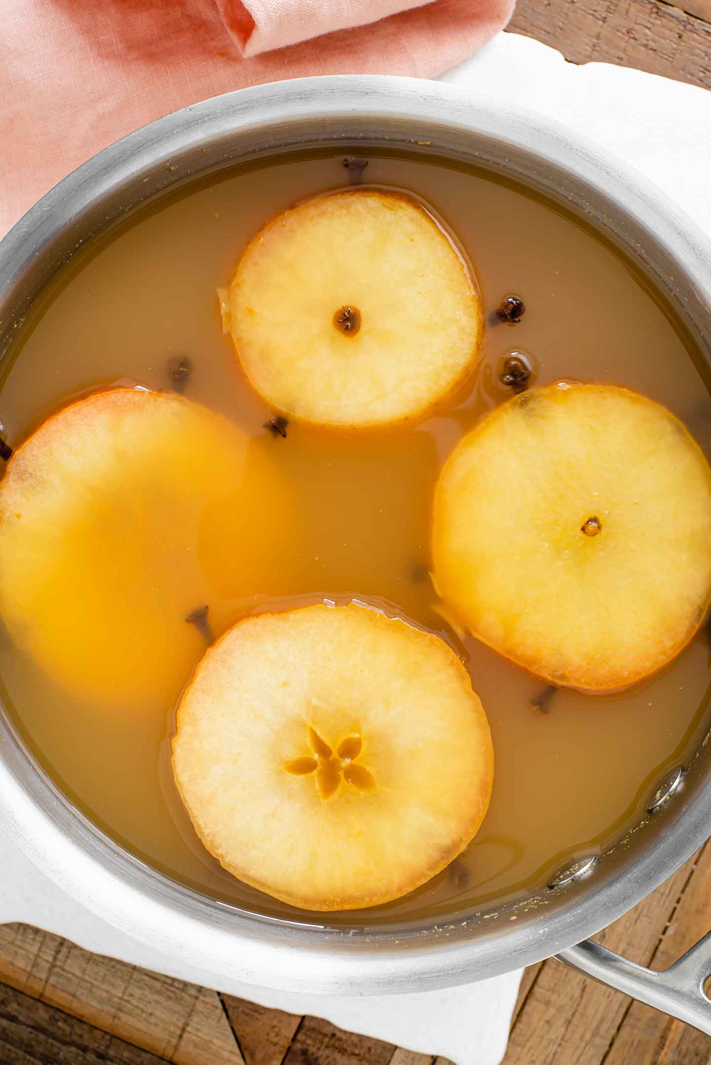 Top down view of hot wassail in a large pot. Four apple rings poked with whole cloves float on the top of the golden liquid.