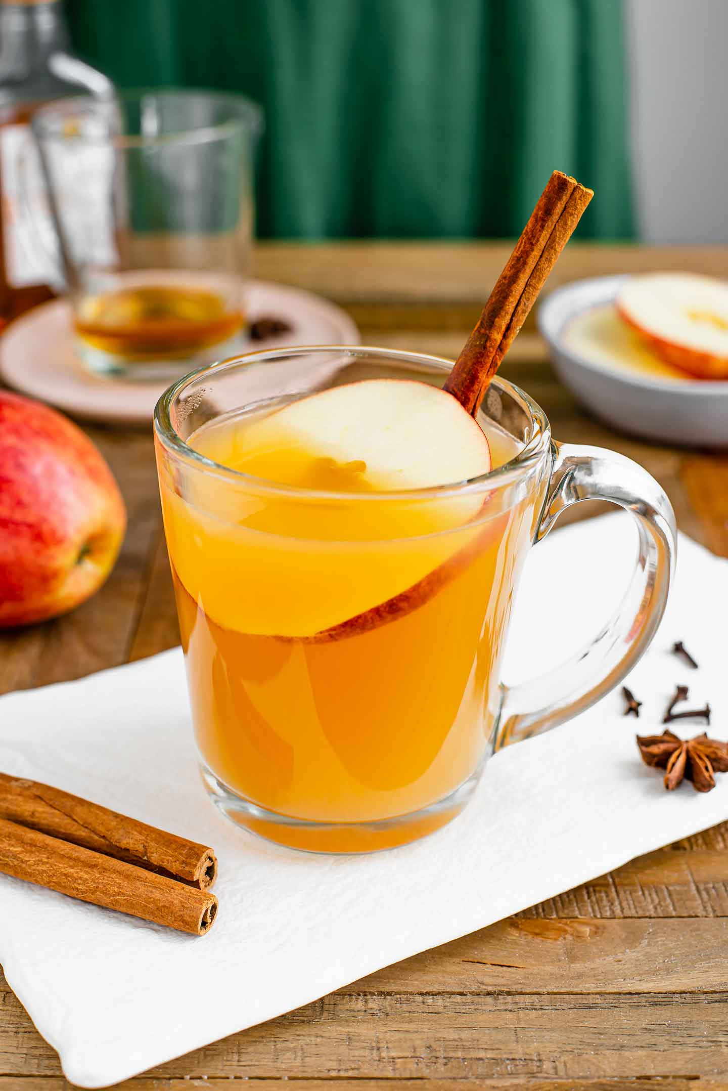 Side view of a glass mug filled with a golden hot wassail. A mug in the background has a small amount of whisky in the bottom and cinnamon sticks, apple slices, and whole cloves are scattered around the mugs.