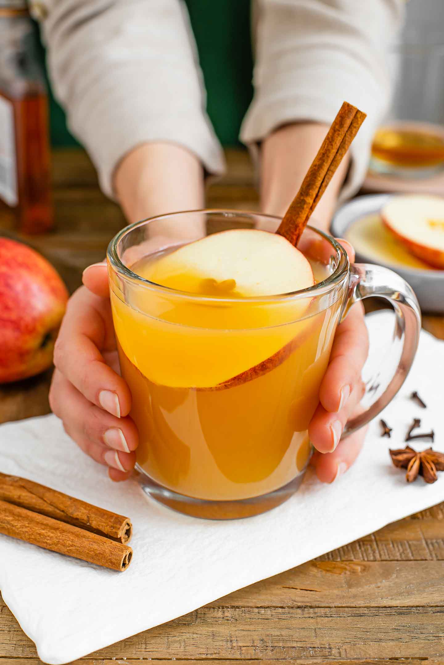 Side view of hands wrapped around a warm glass mug of hot apple cider wassail. A slice of apple and a cinnamon stick garnish the drink.