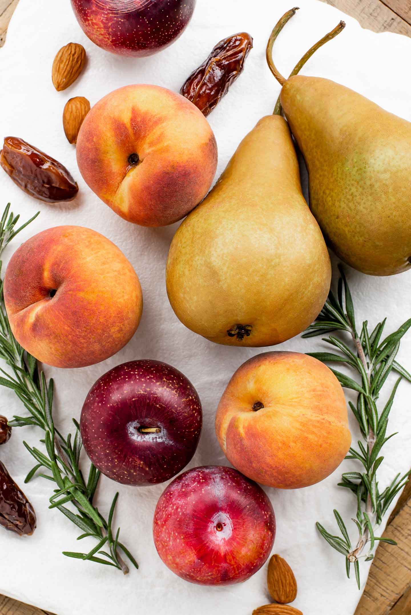 Top down view of pears, peaches, red plums, dates, and sprigs of rosemary scattered on a white tray. 