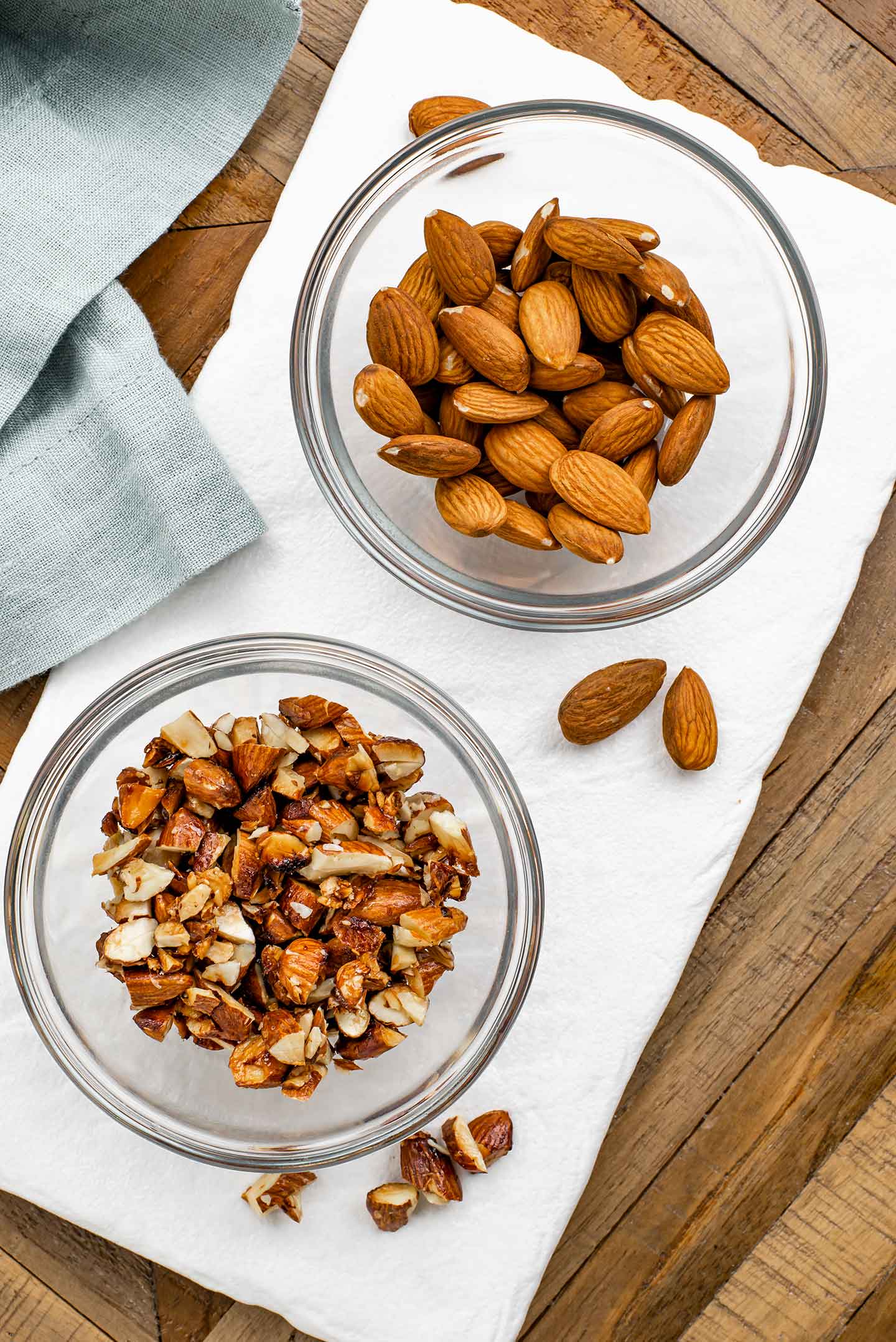 Top down view of two small bowls of almonds on a white tray. In one dish are whole raw almonds. In the other, are coarsely chopped and lightly candied toasted almonds.