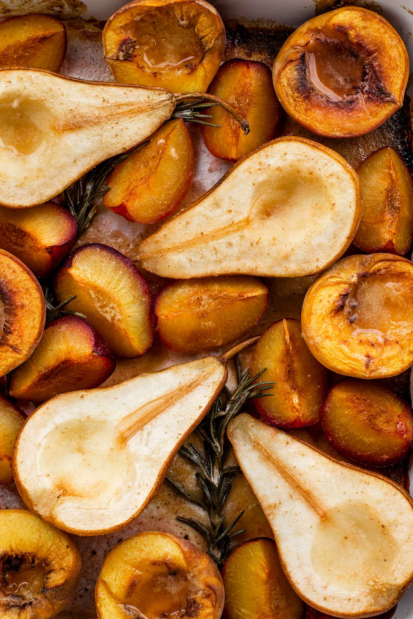Top down view of golden baked pear and stone fruits in a casserole dish with sprigs of rosemary and a bubbly golden syrup.