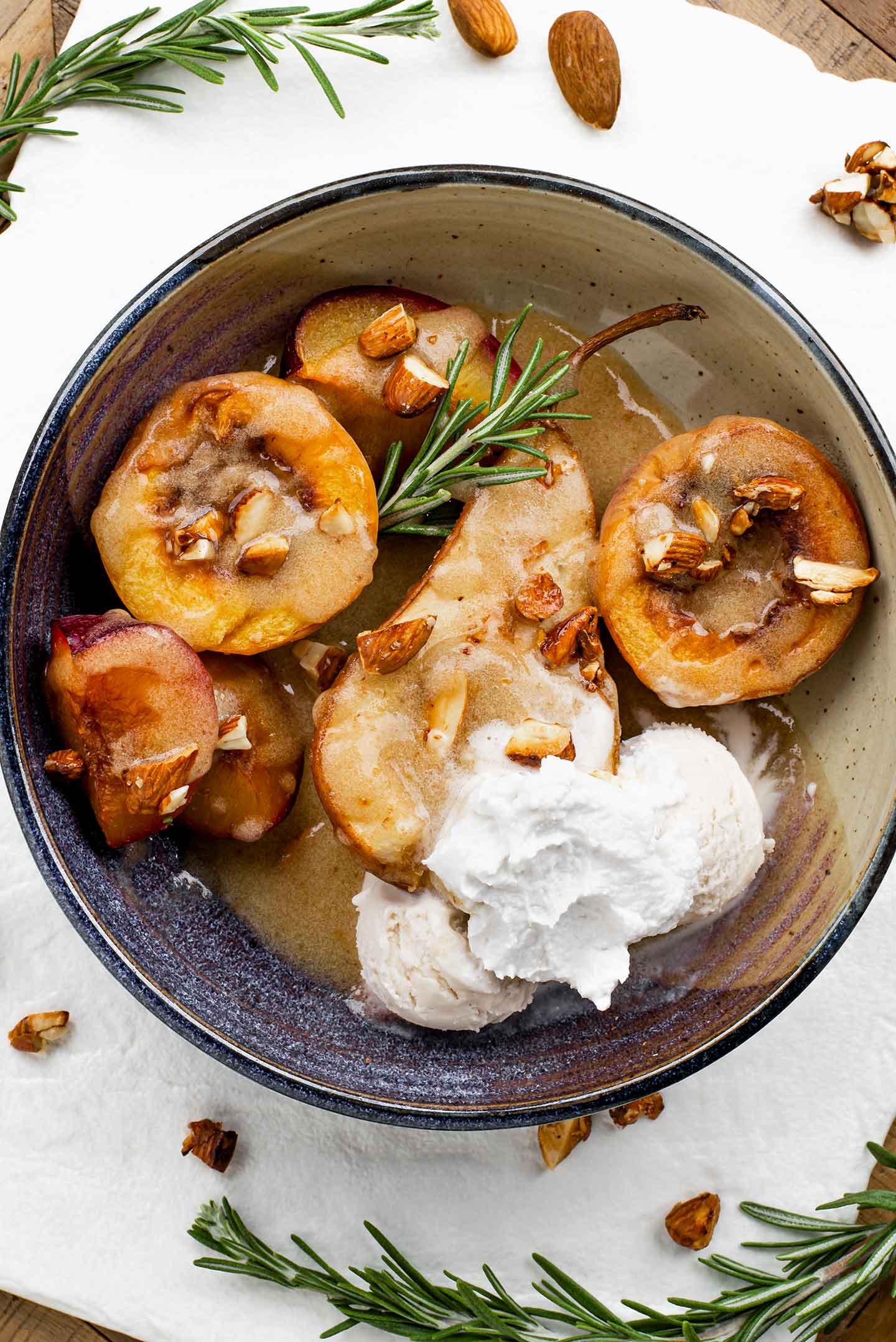 Top down view of a dish with a golden baked pear and stone fruit dessert, drizzled with syrup, garnished with rosemary, and accompanied by a scoop of vanilla ice cream and whipped cream.