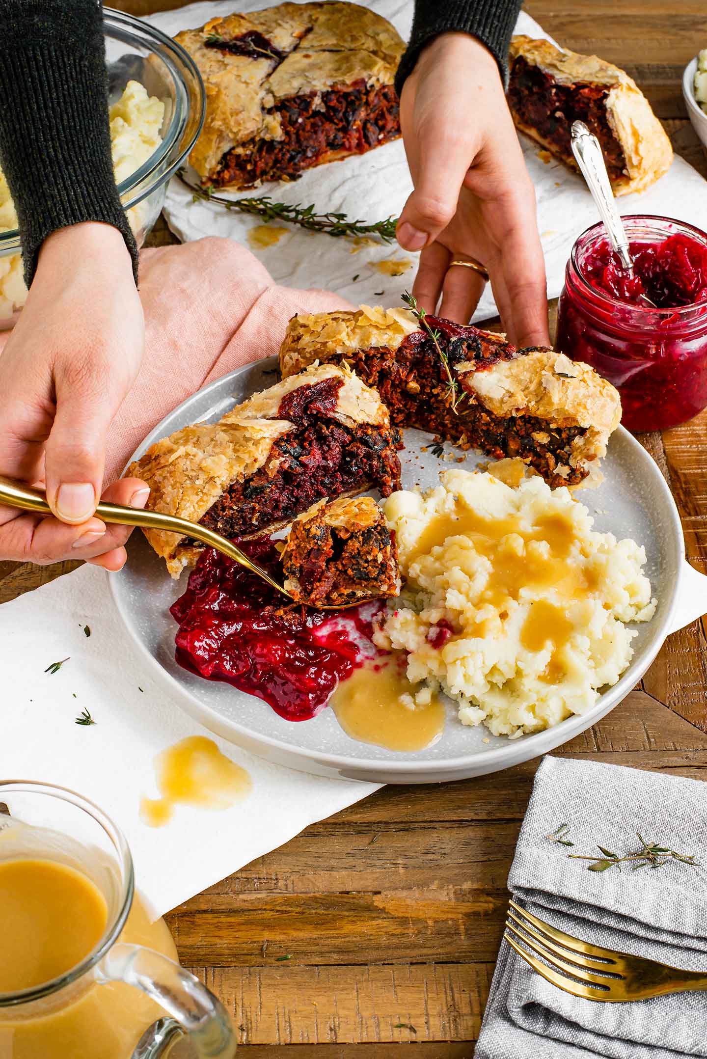 Top down view of two slices of beet wellington on a plate with mashed potatoes, gravy, and cranberry sauce. A hand holds up a fork with a slice of beet wellington.