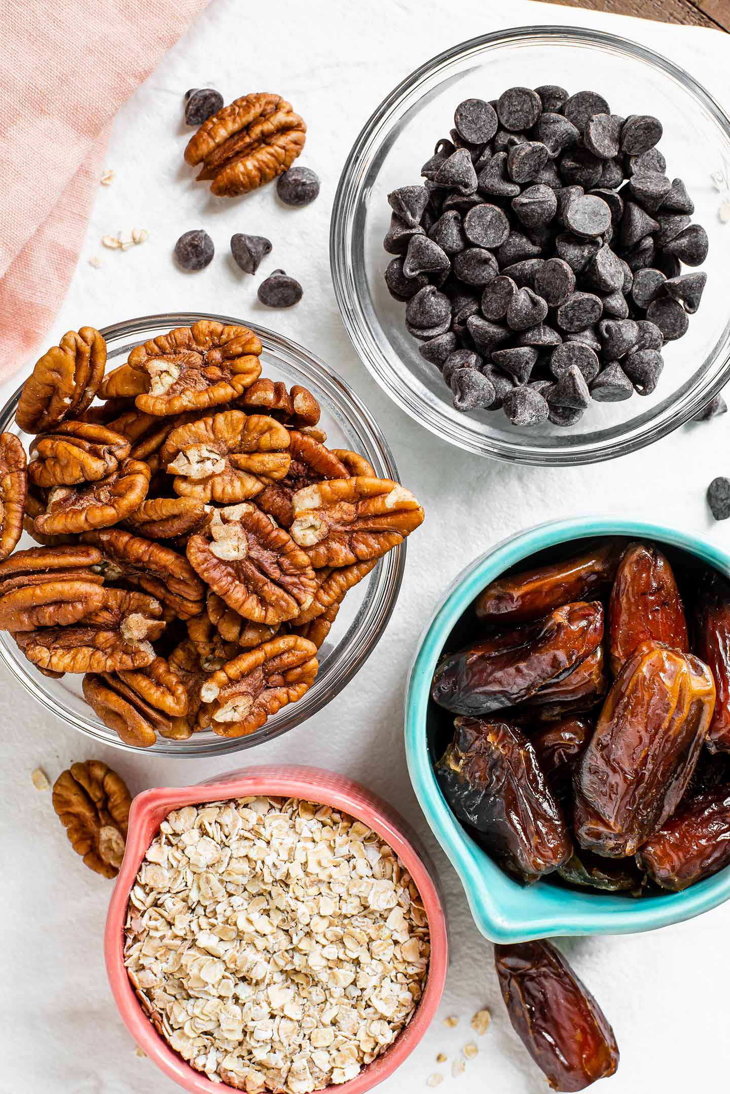 Top down view of ingredients on a white tray. Pitted dates, pecan halves, chocolate chips, and oats fill separate bowls.