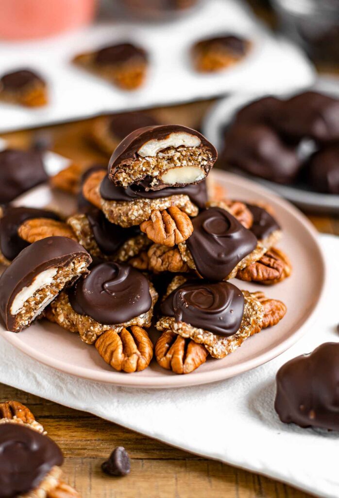 Side view of date chocolate turtles piled high on a plate. The turtle on the very top of the stack is sliced in half revealing the pecan and date caramel filling.