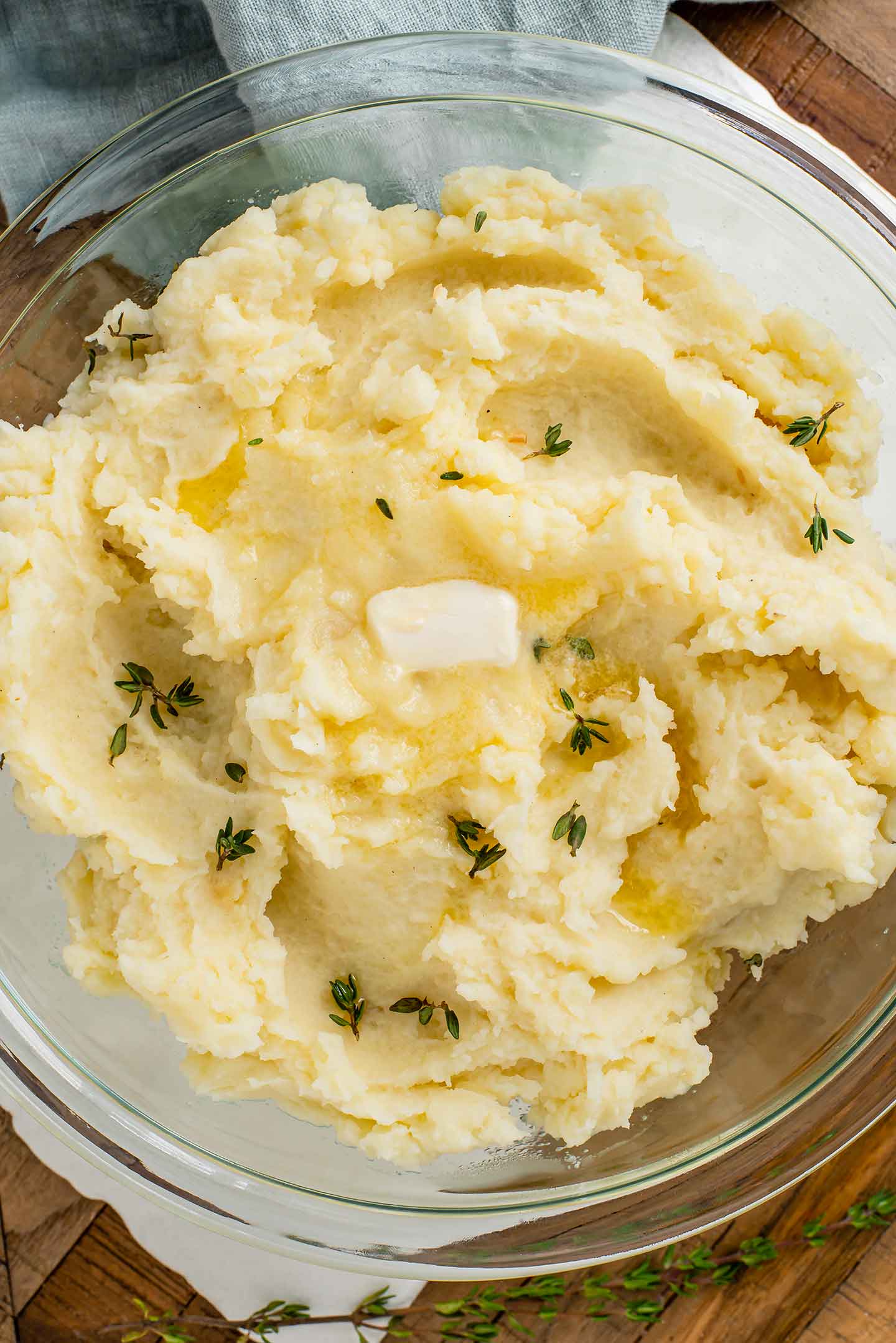 Top down view of fluffy mashed potatoes in a glass bowl. Fresh thyme is sprinkled on the potatoes and a slice of vegan butter melts on the top.