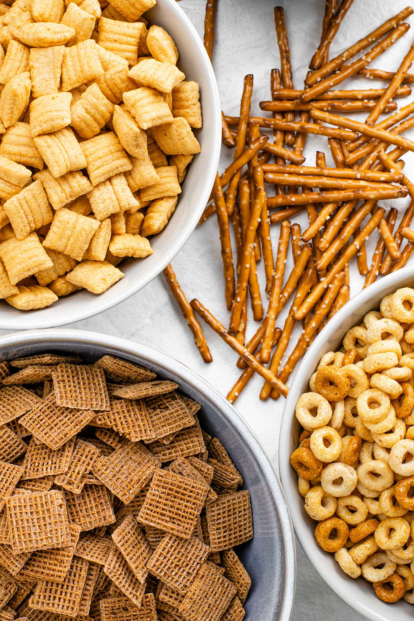 Top down view of cheerios, shreddies, corn squares, and pretzel sticks on a white tray.