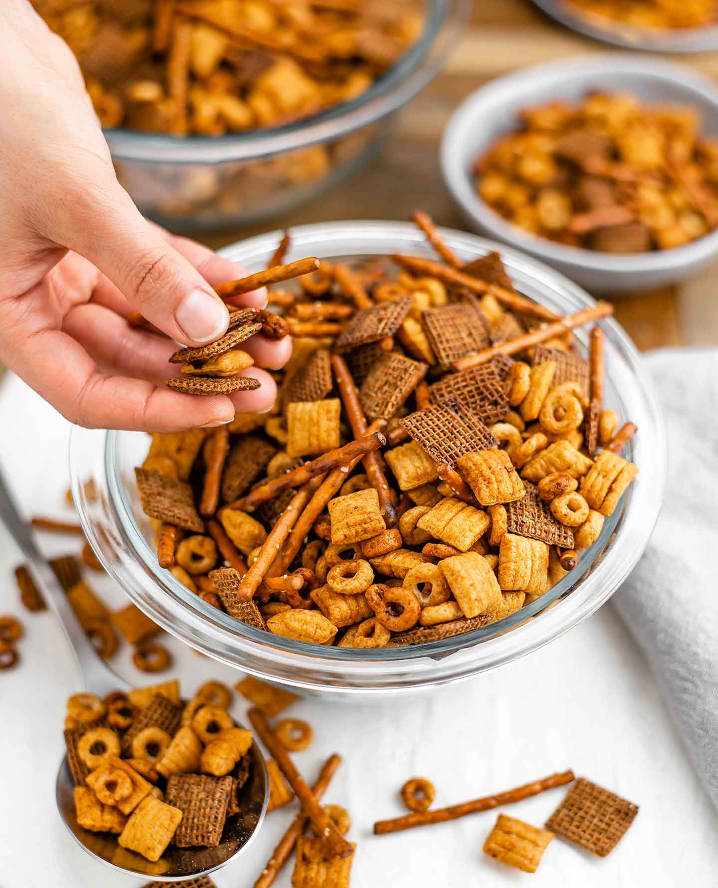 Side view of hand holding pretzels sticks and pieces of cereal from an overflowing bowl of toasty vegan party mix.