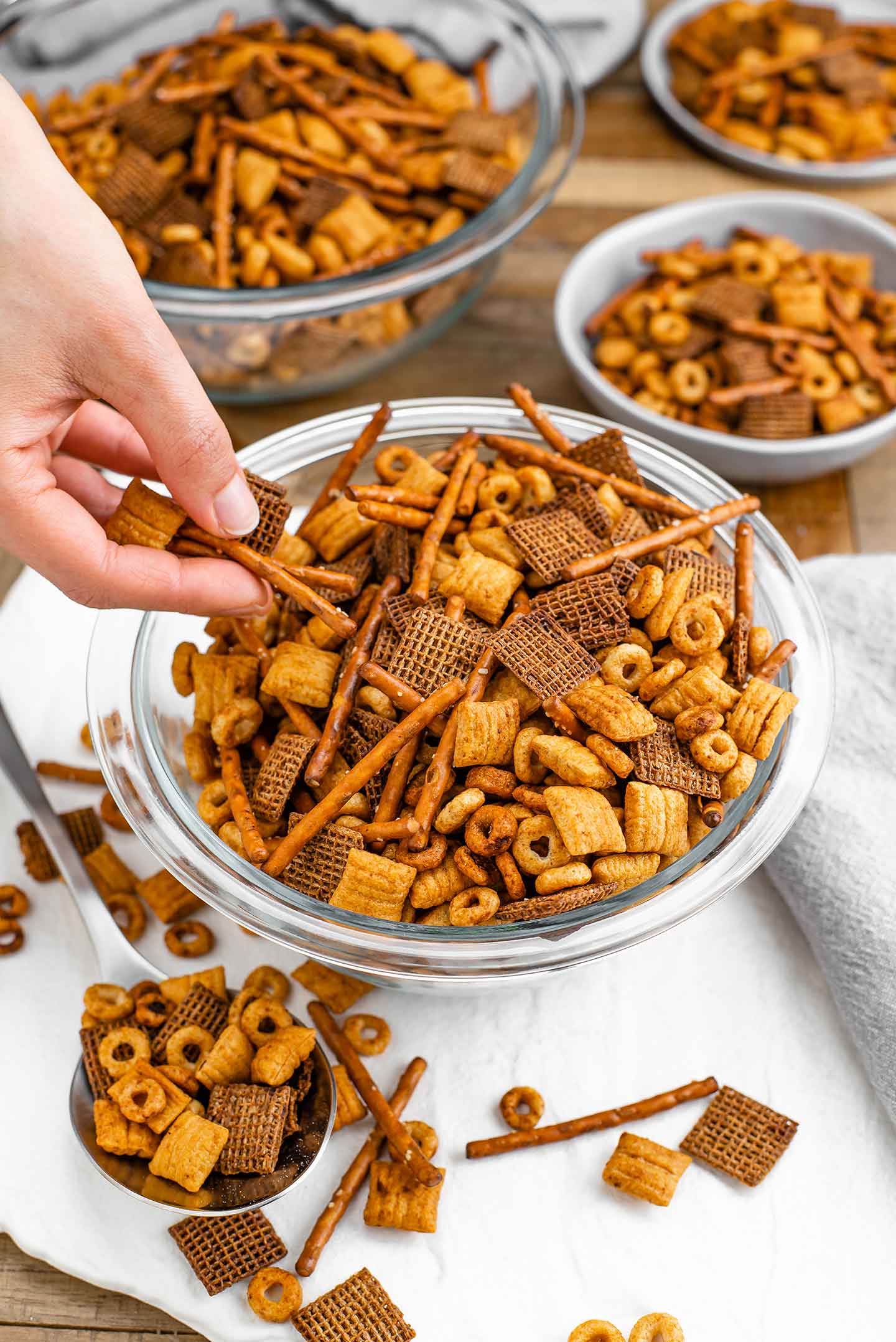 Side view of hand grabbing party mix from a large bowl of a spiced pretzel and cereal mixture. Smaller bowls of party mix are in the background.