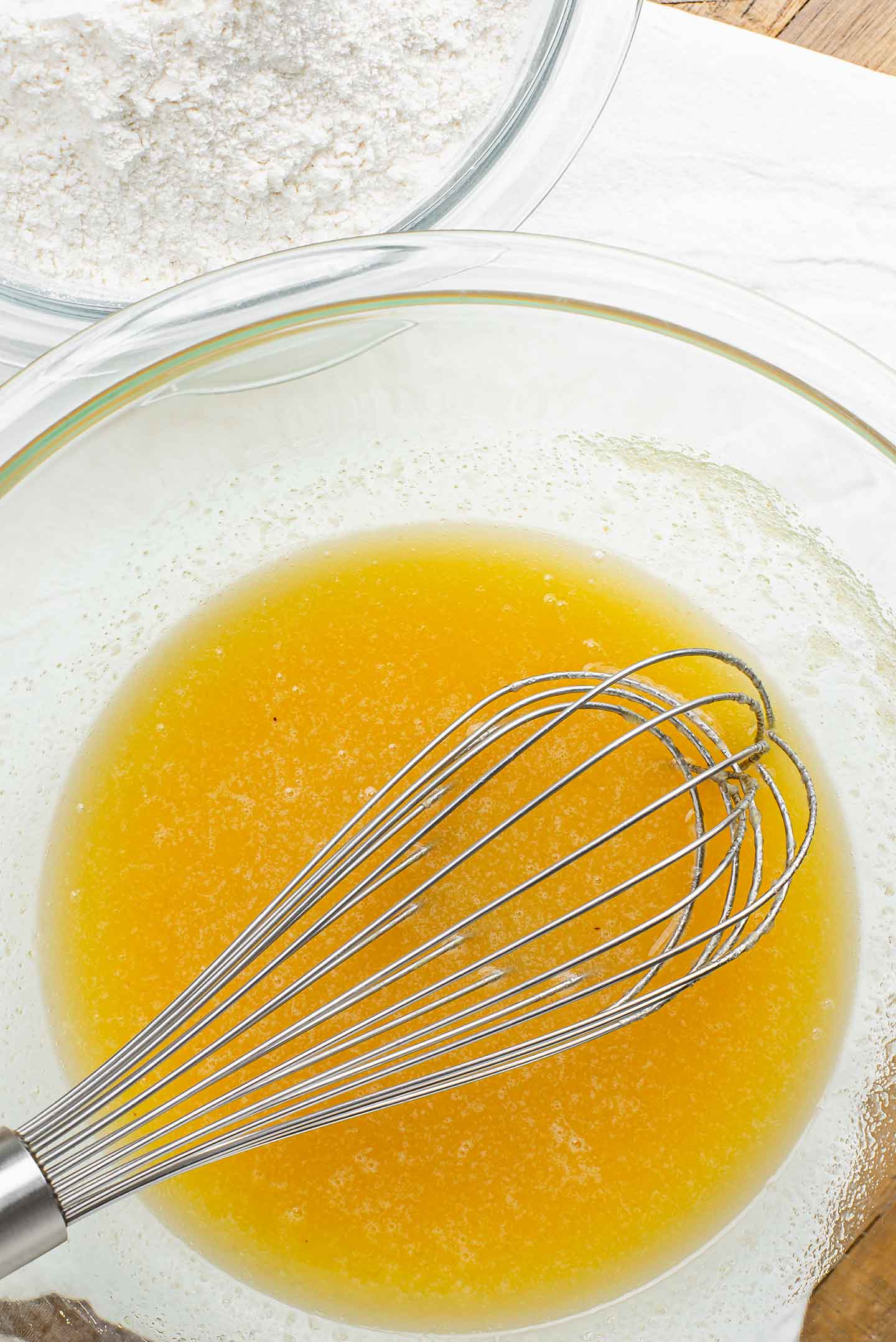 Top down view of a glass bowl with a stirred mixture of applesauce, melted vegan butter, and sugar. A metal whisk rests in the bowl.