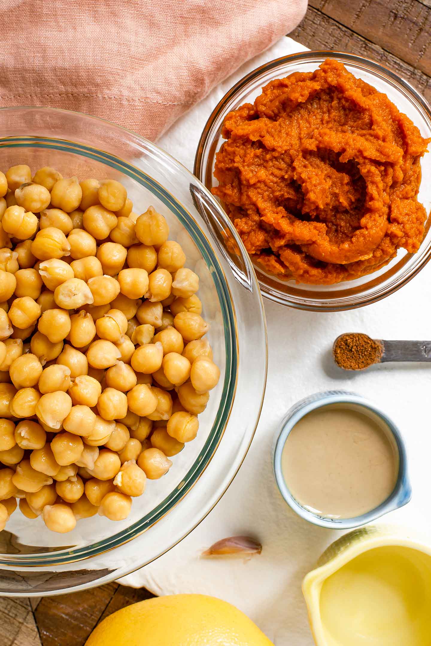 Top down view of ingredients on a white tray. A bowl of cooked chickpeas rests beside a small bowl of pumpkin puree, a dish of tahini, lemon juice, vinegar, a garlic clove, and a spoon of nutmeg.