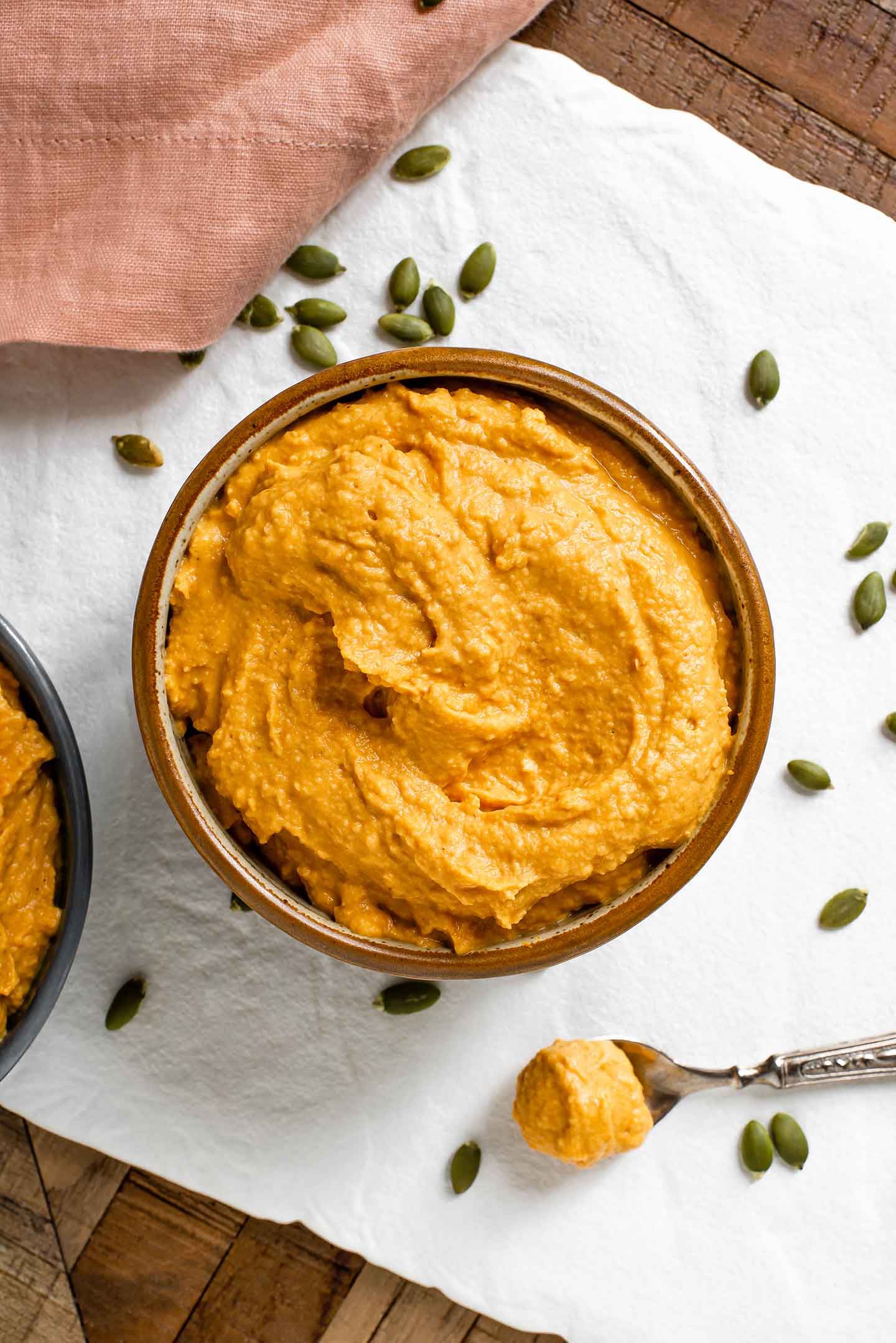 Top down view of pumpkin hummus filling a ceramic bowl. A small spoon with a scoop of the hummus rests on a whit tray sprinkled with pumpkin seeds. 