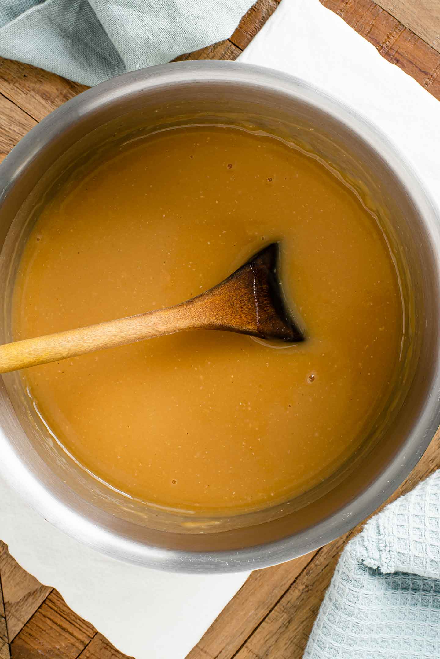 Top down view of a golden coloured gravy in a pot with a wooden spoon stirring the gravy.