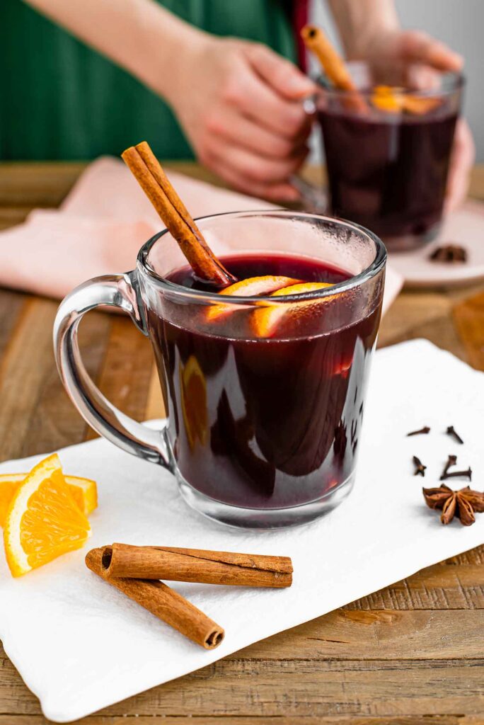 Side view of a mug of mulled wine on a white tray. Cinnamon sticks, orange slices, and whole cloves decorate the tray and a cinnamon stick rests in the hot wine.