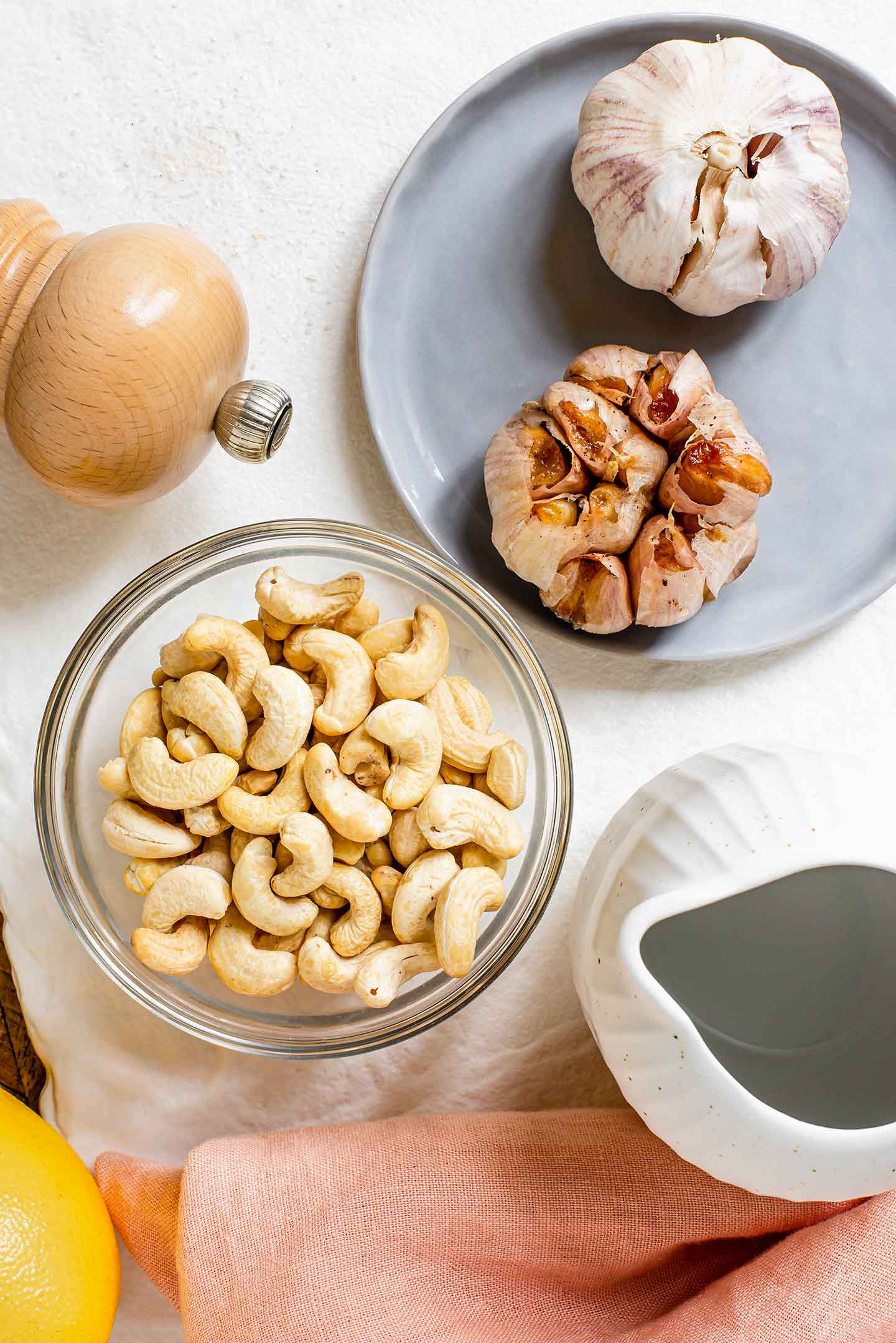 Top down view of ingredients. A bulb of roasted garlic rests beside an raw bulb of garlic. A small dish of raw cashews is surrounded by a lemon, a salt mill and a small jug of water. 