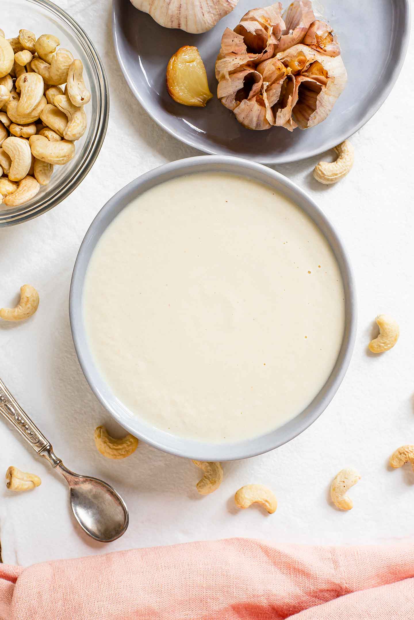 Top down view of the creamy white sauce in a shallow dish surrounded by raw cashews and roasted garlic.