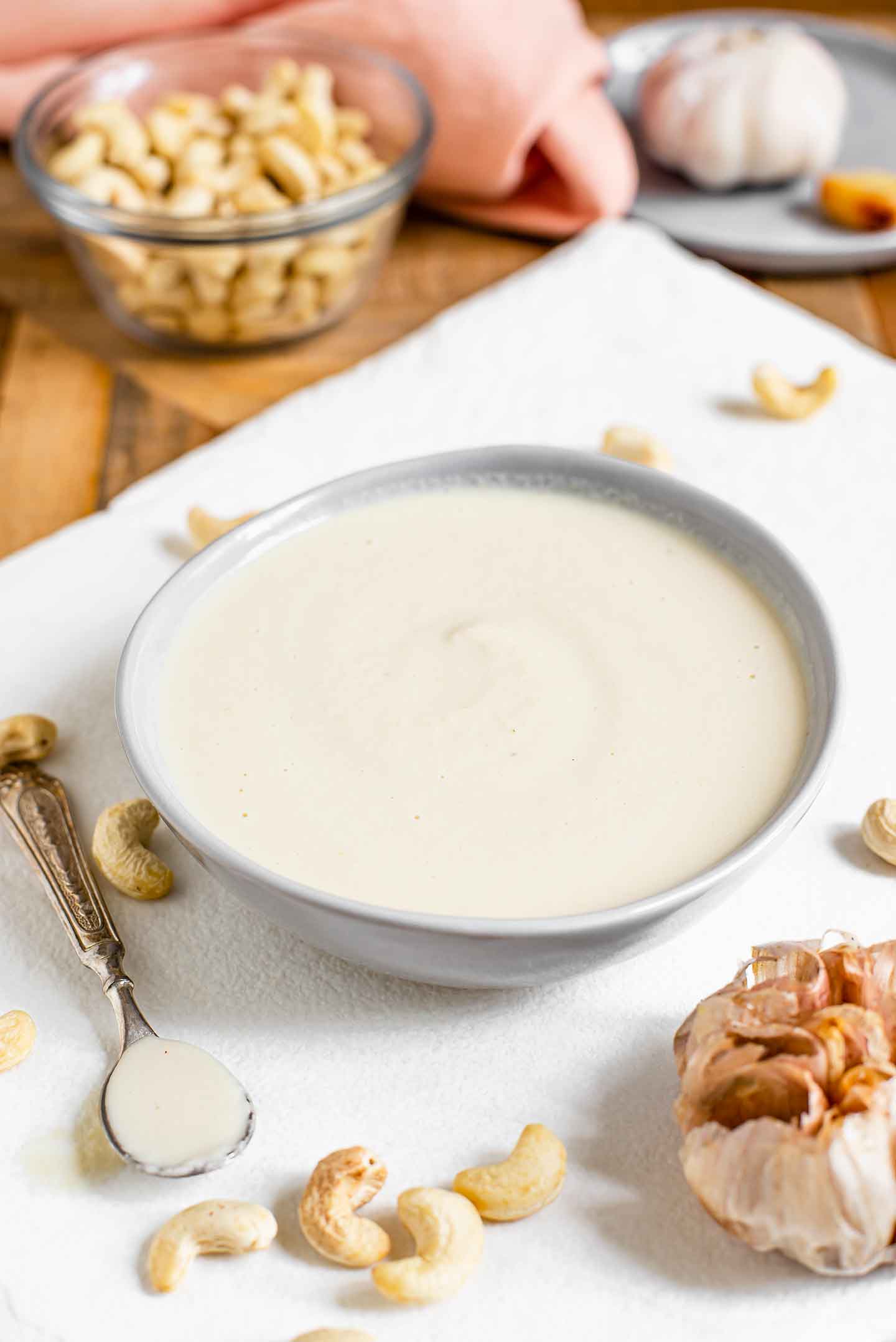 Side view of a roasted garlic cashew cream sauce filling a small dish. A spoon of the creamy sauce rests beside the bowl on a white tray garnished with raw cashews and a bulb of roasted garlic. 