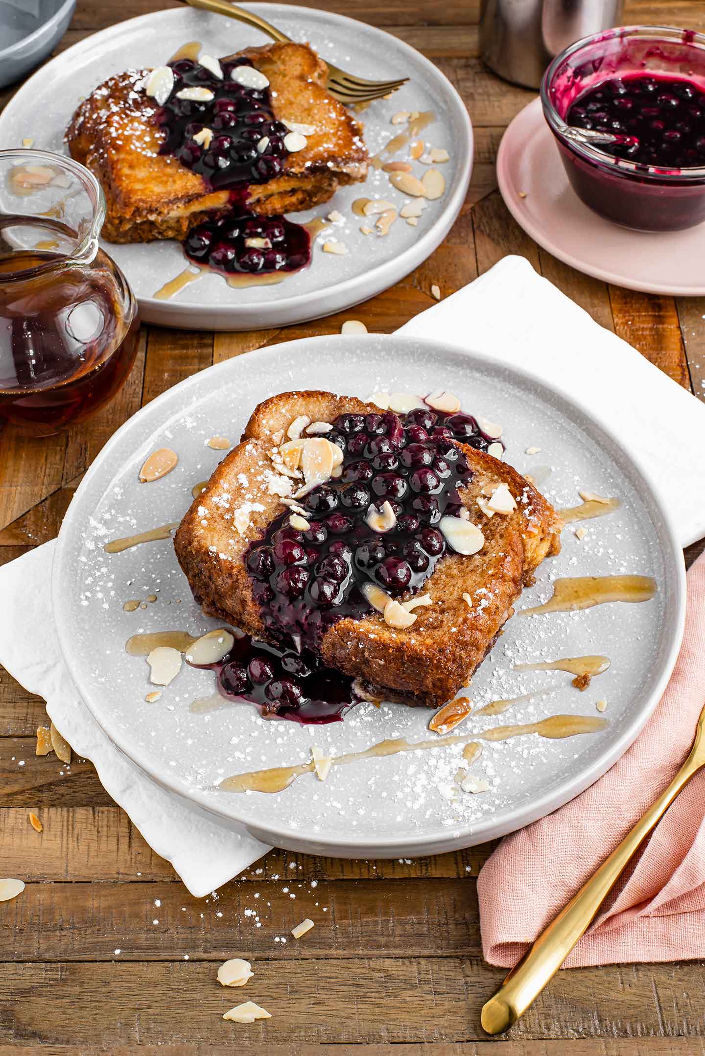 Top down view of two plates of vegan french toast casserole topped with blueberry sauce, maple syrup, toasted almonds, and powdered sugar.