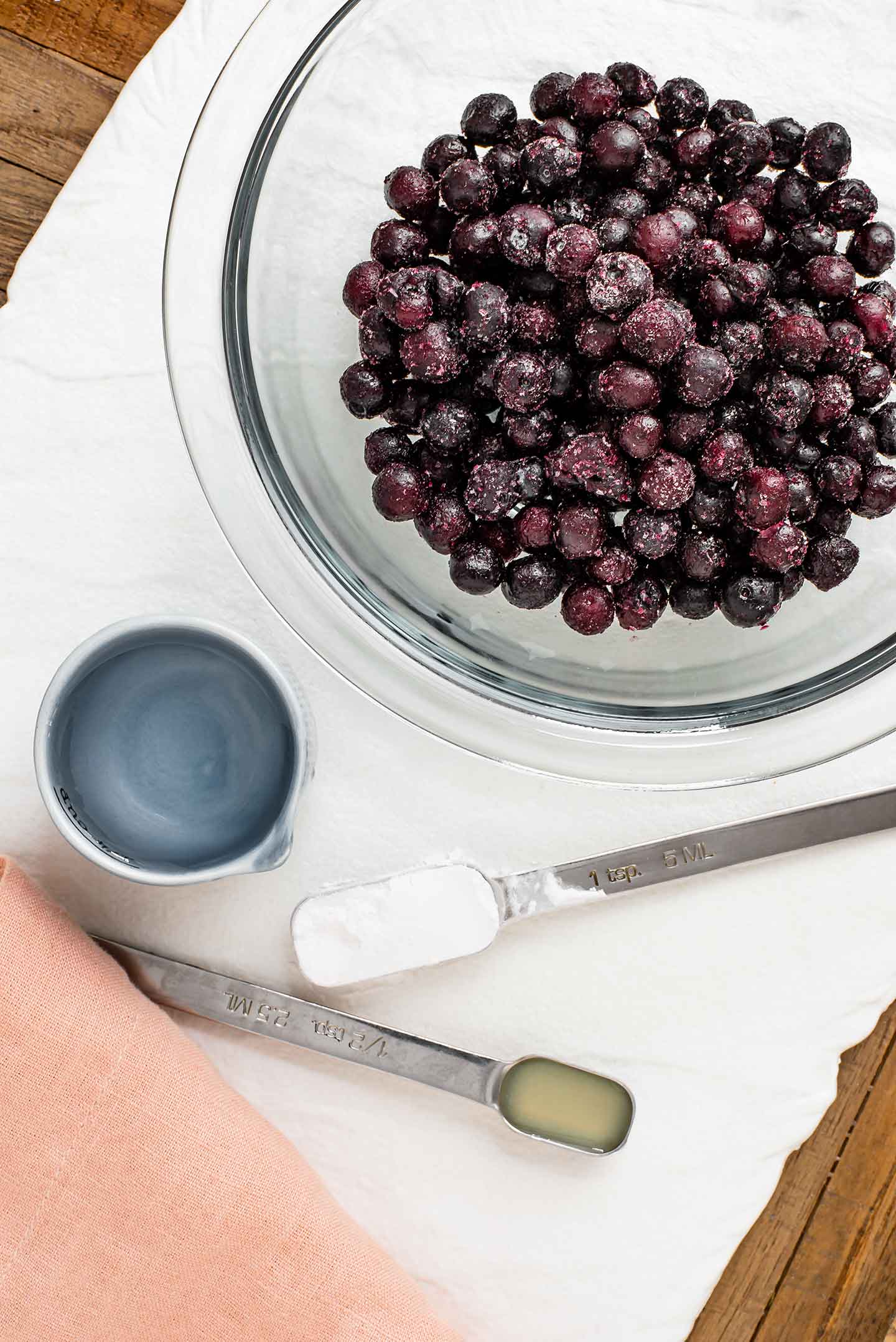 Top down view of frozen blueberries in a glass bowl on a white tray. A measuring spoon of starch and a small dish of water sit next to the berries.
