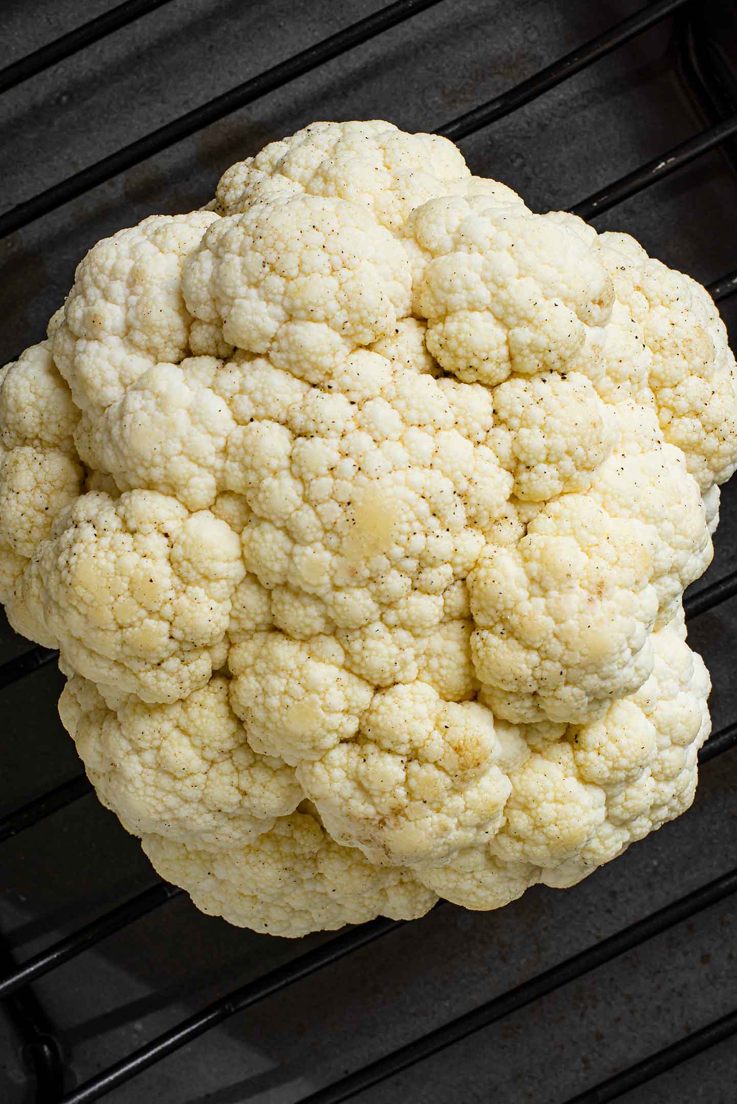 Top down view of a trimmed cauliflower, basted in oil, and placed on the rack of a roasting tray.
