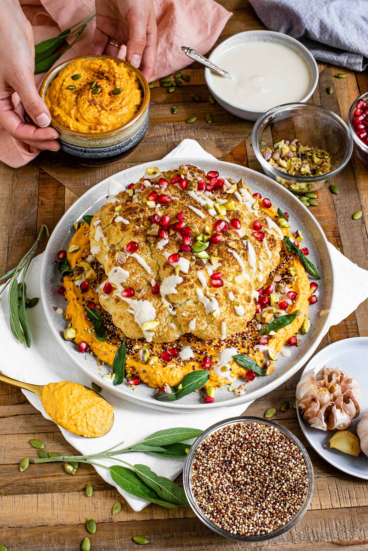 Top down view of a golden cauliflower atop an orange bed of pumpkin hummus, sprinkled with popped quinoa, drizzled with a cream sauce, and garnished with pomegranate, pistachios, and sage leaves.
