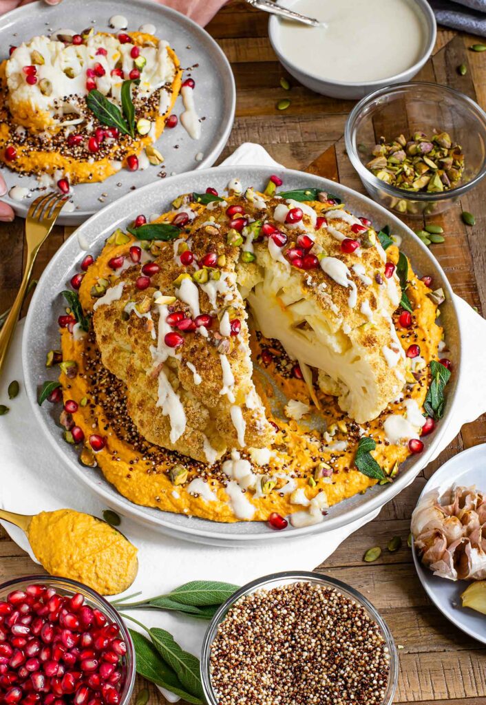 Top down view of a large slice removed from a beautifully garnished whole roasted cauliflower. An individual serving rests on a small plate.