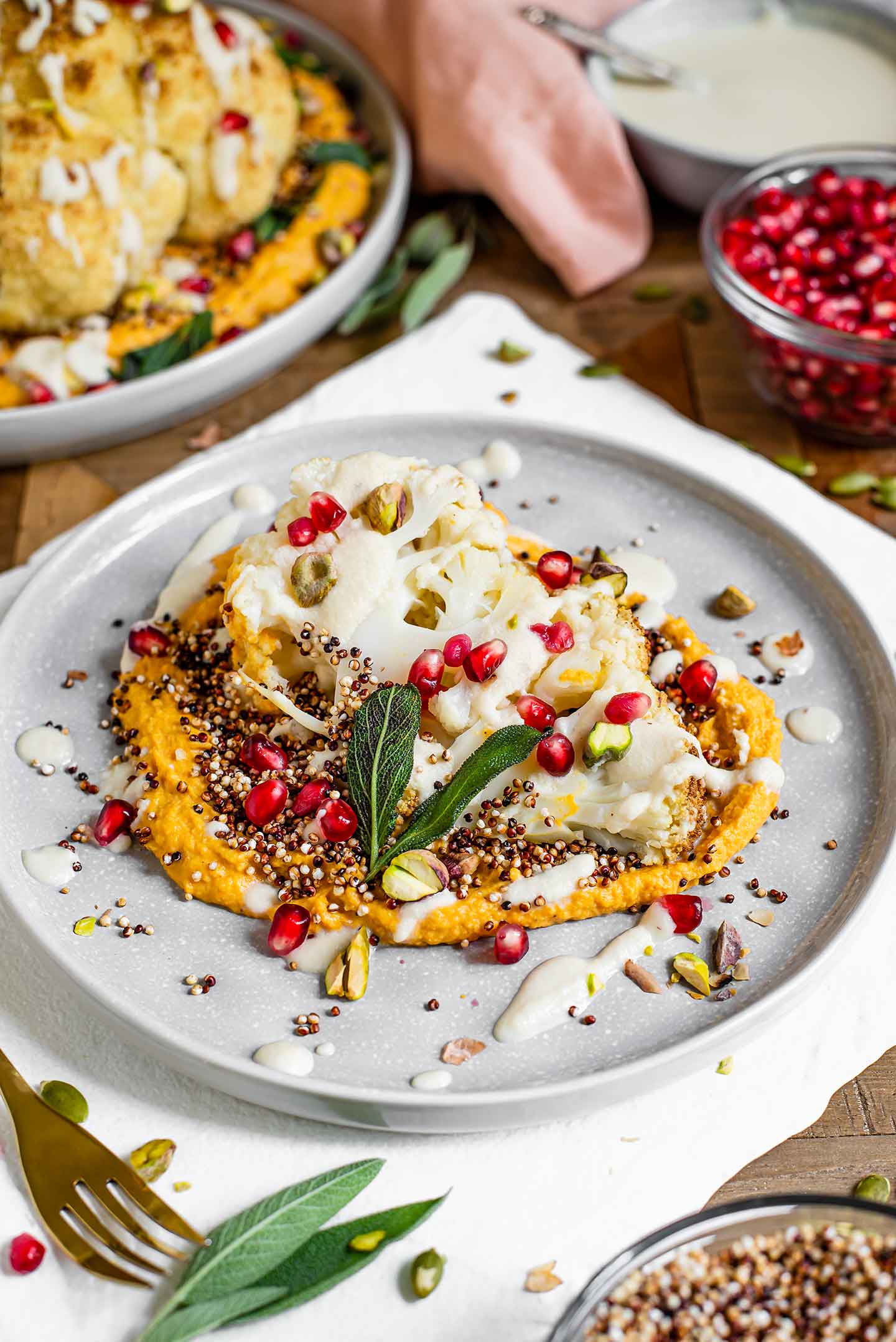 Side view of a small plate with a sliced cauliflower steak. The steak sits on a bed of hummus and is garnished with popped quinoa, pomegranate, pistachios, a creamy sauce, and crispy sage leaves.