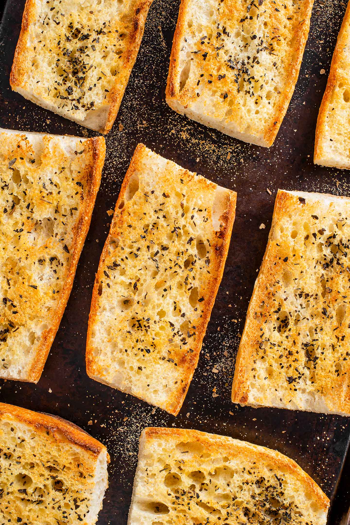 Top down view of toasted garlic bread on a baking sheet. The bread is golden with melted butter and sprinkled with garlic powder and dried herbs.