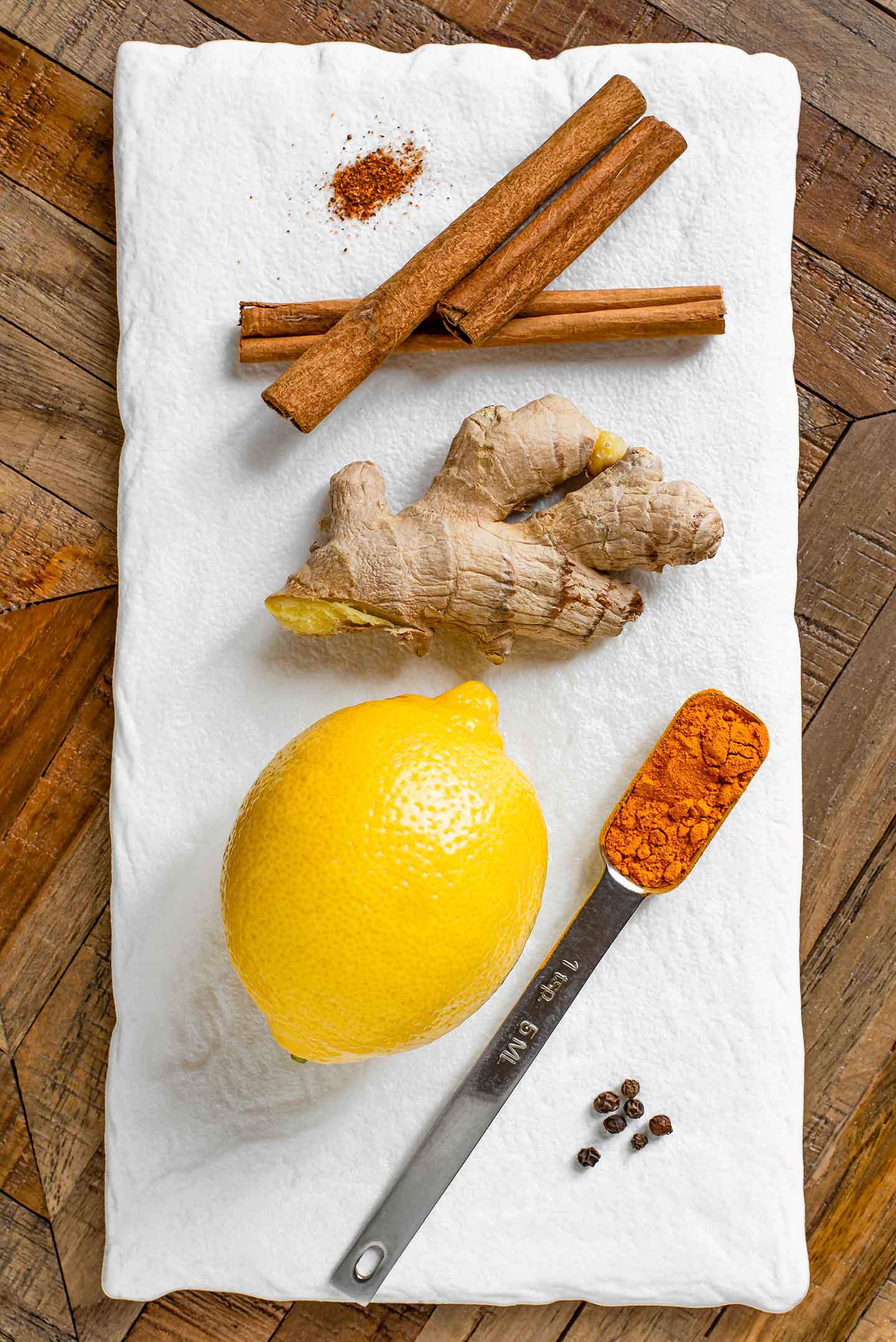 Top down view of ingredients. Fresh ginger, a lemon, cinnamon sticks, ground turmeric, black peppercorns, and ground cayenne pepper are displayed on a white tray.