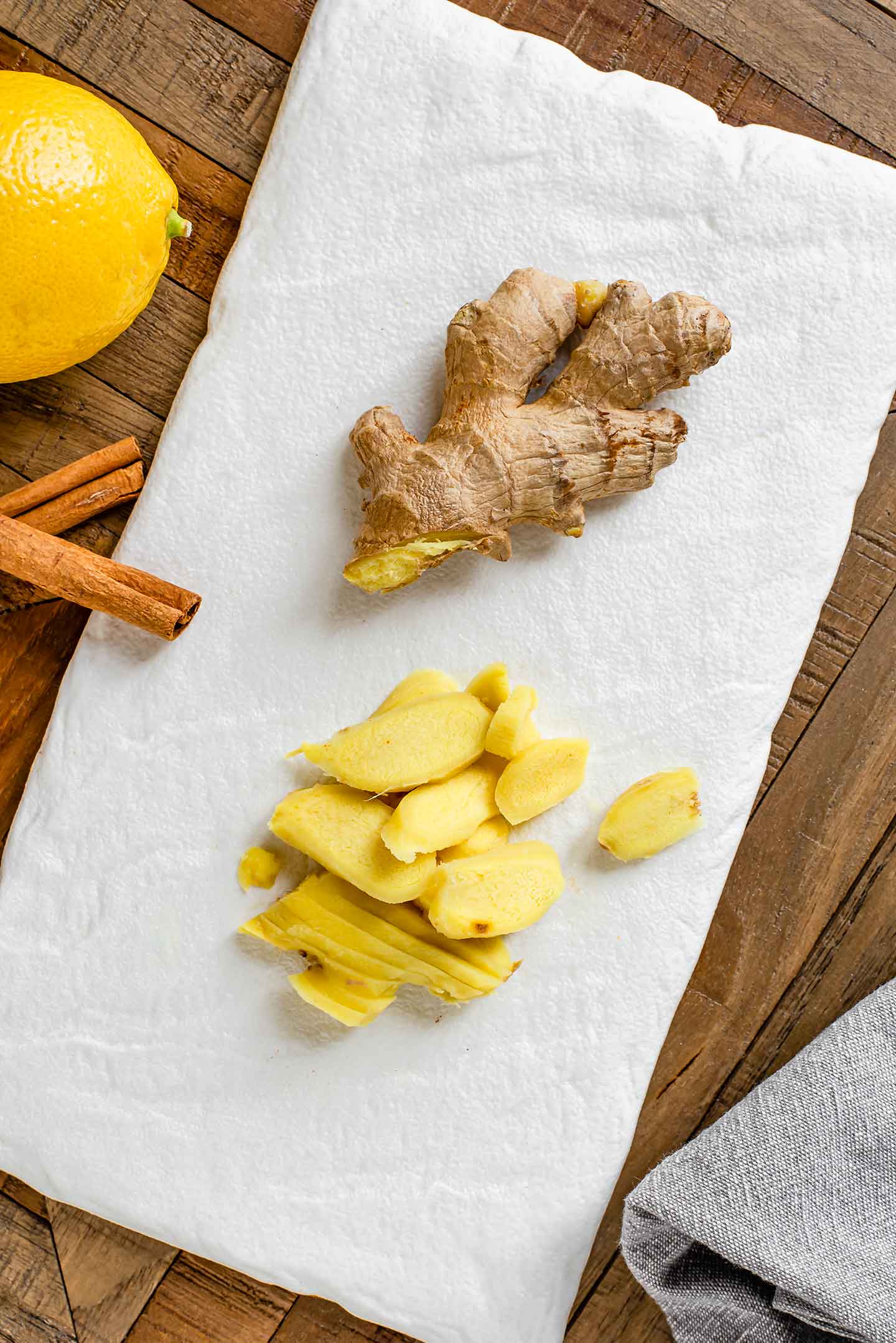 Top down view of peeled and sliced ginger root on a white tray with cinnamon sticks and a lemon.