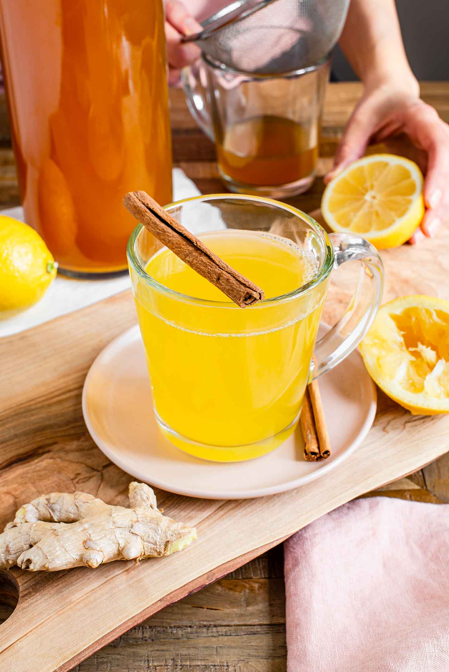 Side view of a bright yellow tea in a glass mug. Fresh ginger root and lemon surround the mug. A cinnamon stick rests on the rim.