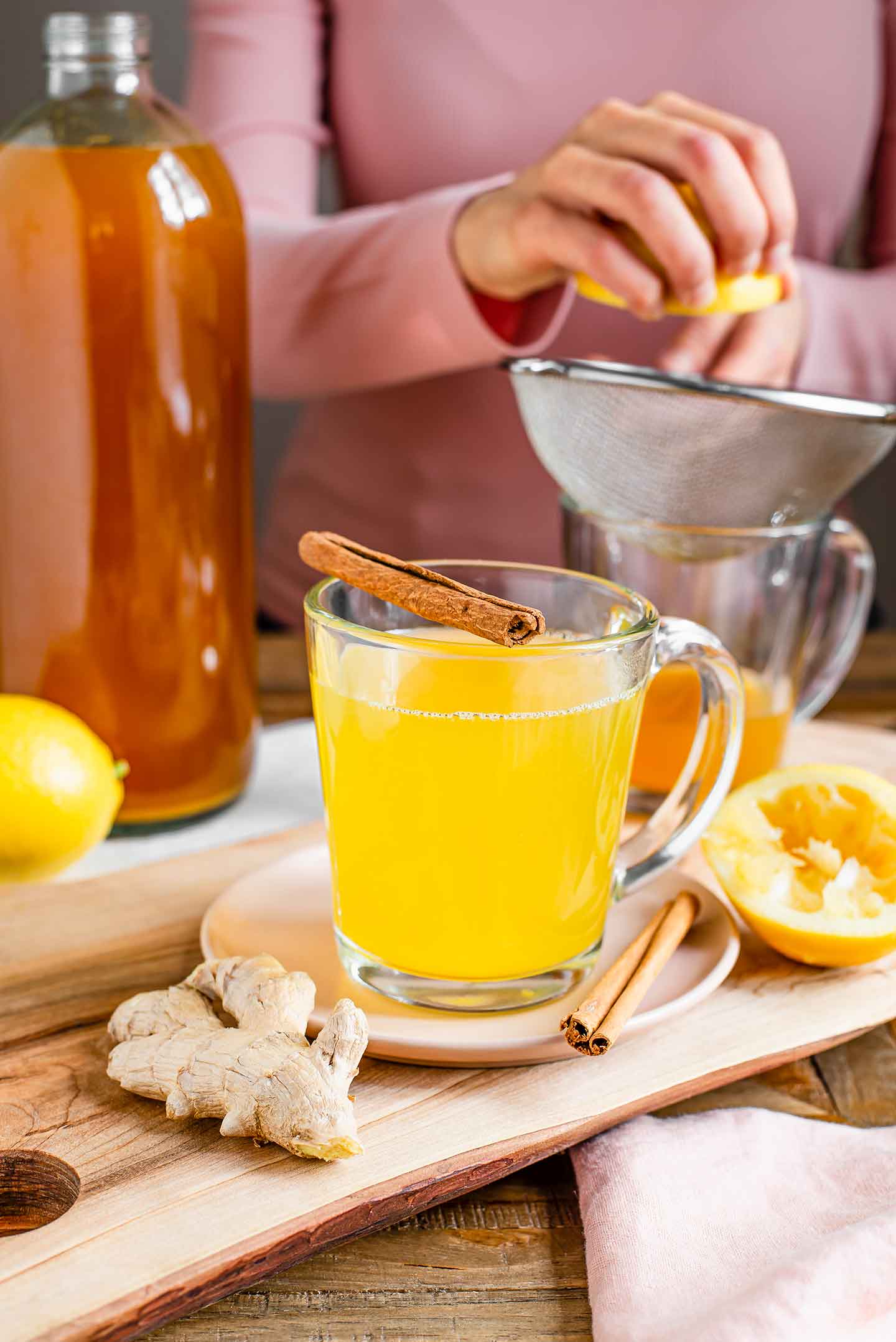 Side view of a mug of tea in the foreground with a large glass bottle of a darker concentrate in the background. A woman squeezes lemon juice through a strainer into a mug with a small portion of the concentrate.