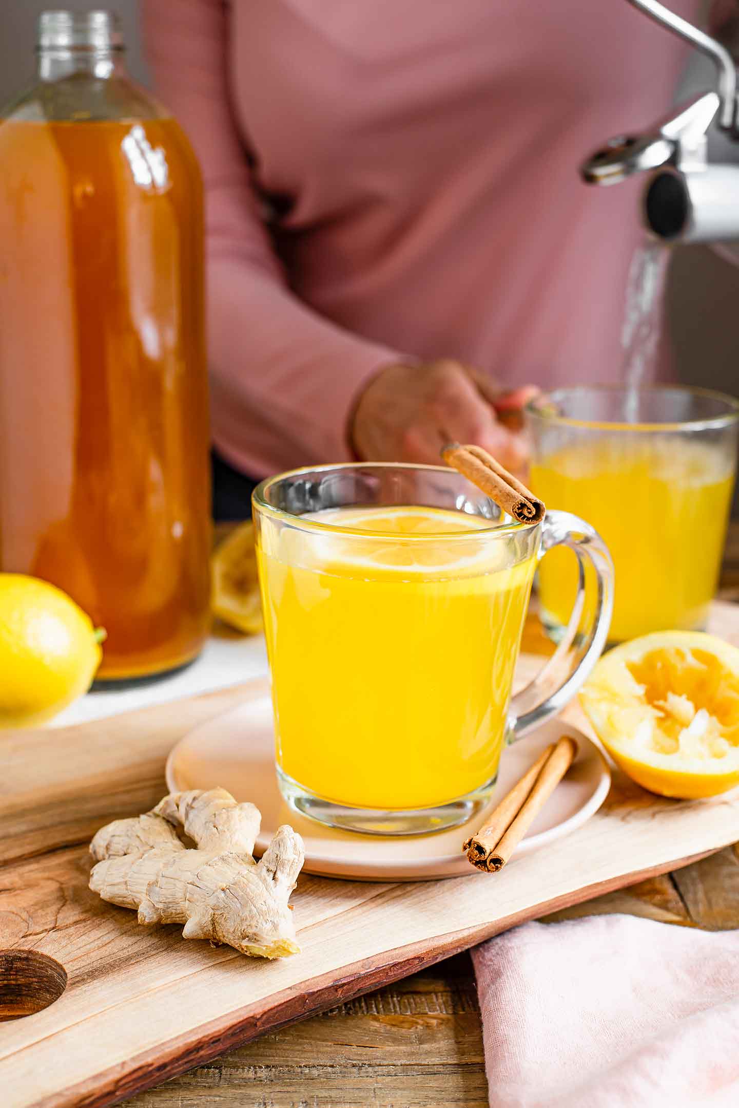 Side view of a mug of ginger turmeric tea. A woman pours hot water from a kettle into a second mug of tea in the background.