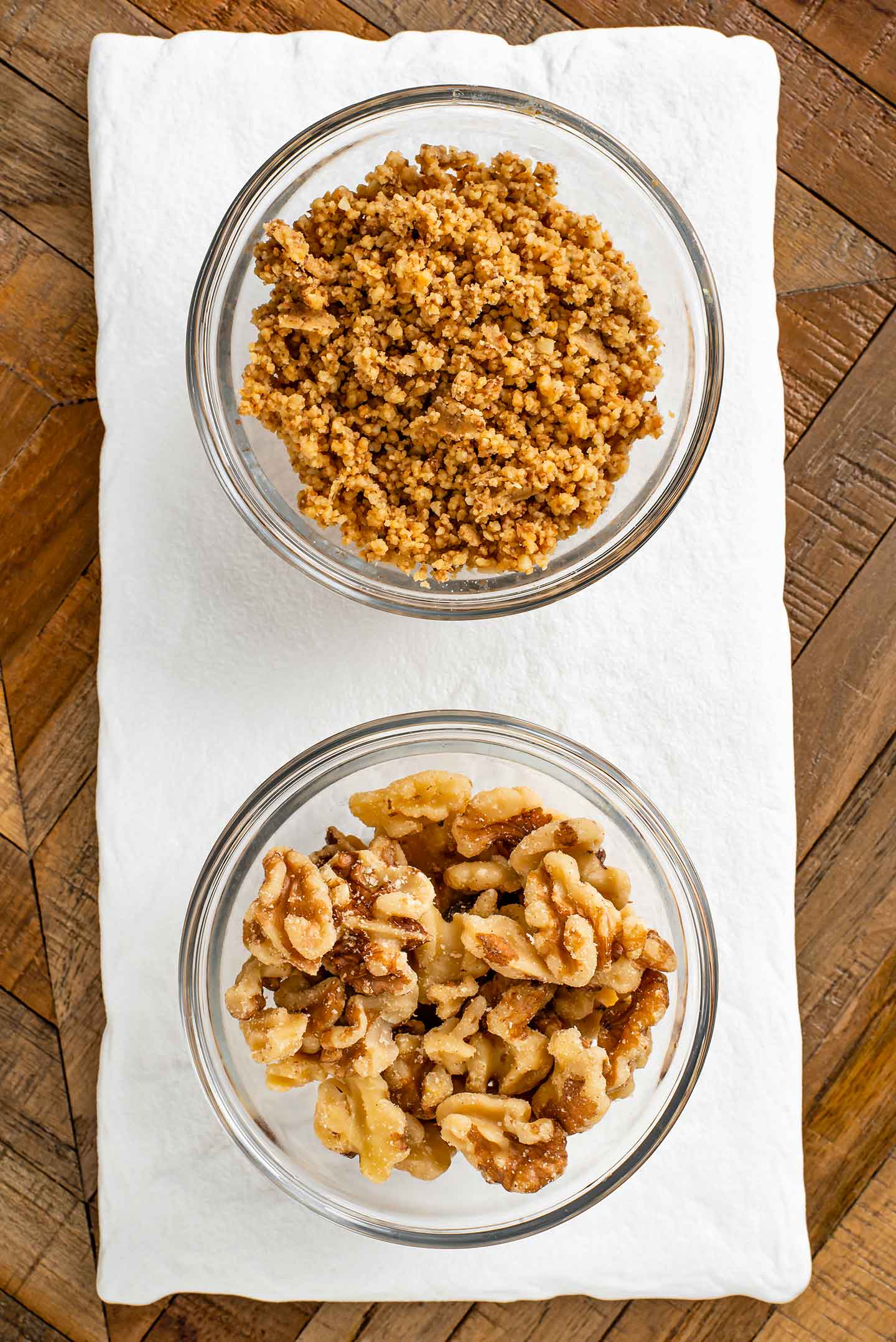 Top down view of two small bowls of walnuts. One bowl has halved raw walnuts and the other has toasted and ground walnuts.